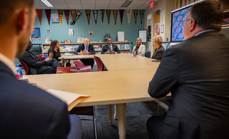 A photograph of adults in suits sitting around a large square made of wooden desks in a school library.