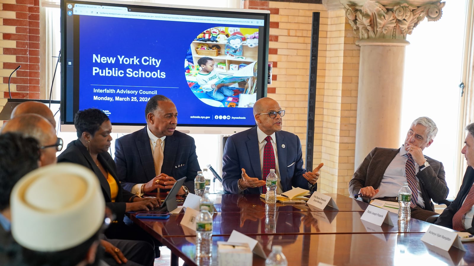 A group of men and women sit around a conference table with a New York City Public Schools slide in the background.