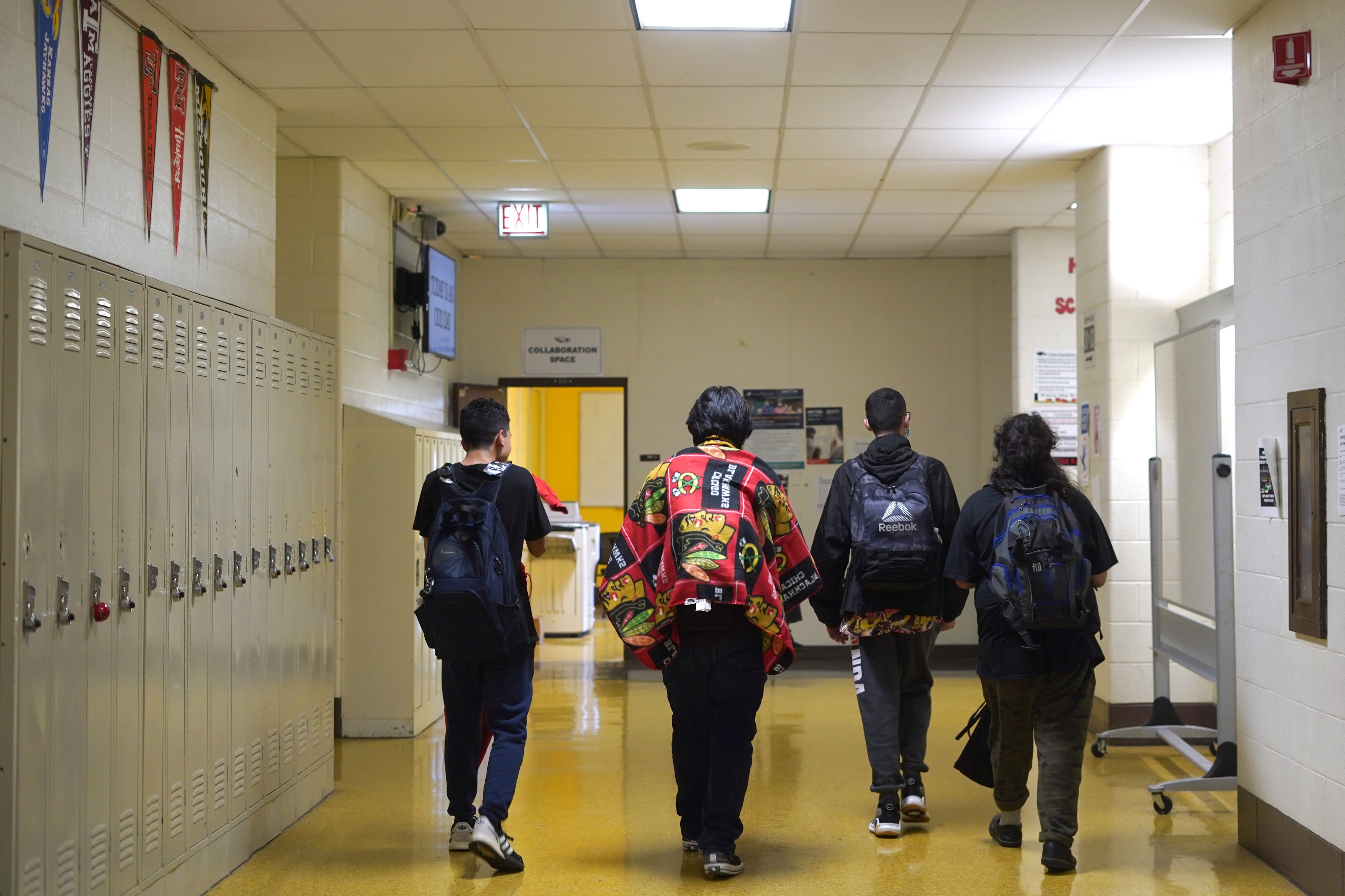 Four students with backpacks walk down a hallway with lockers on the left of them.