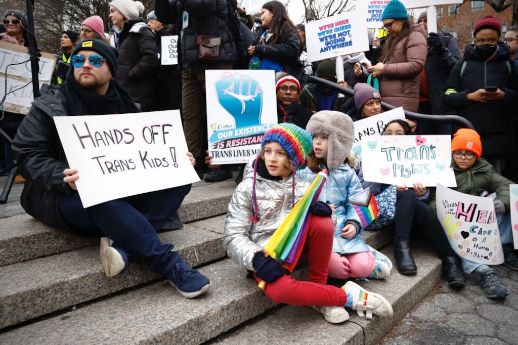 A group of children and adults, bundled up against the cold, sit on stone steps. They hold colorful signs in support of trans rights. "Hands off trans kids," reads one sign.