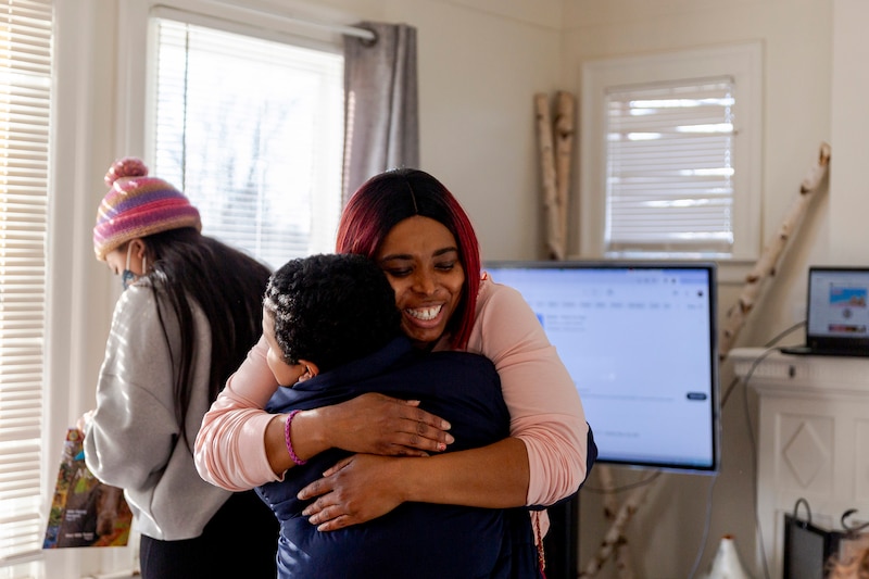 A woman wearing a pink shirt hugs a child in a house with another adult in the background,