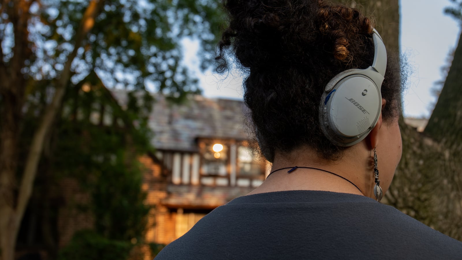 A teen wearing headphones is standing outside in a leafy neighborhood. 