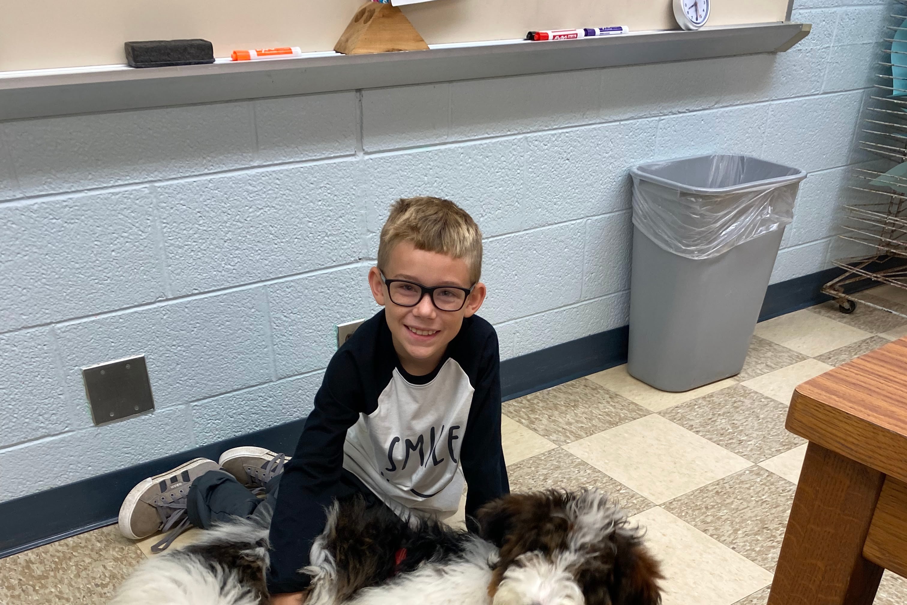 a student sits on the linoleum floor of a classroom with a hand on the dog sprawled in front of him