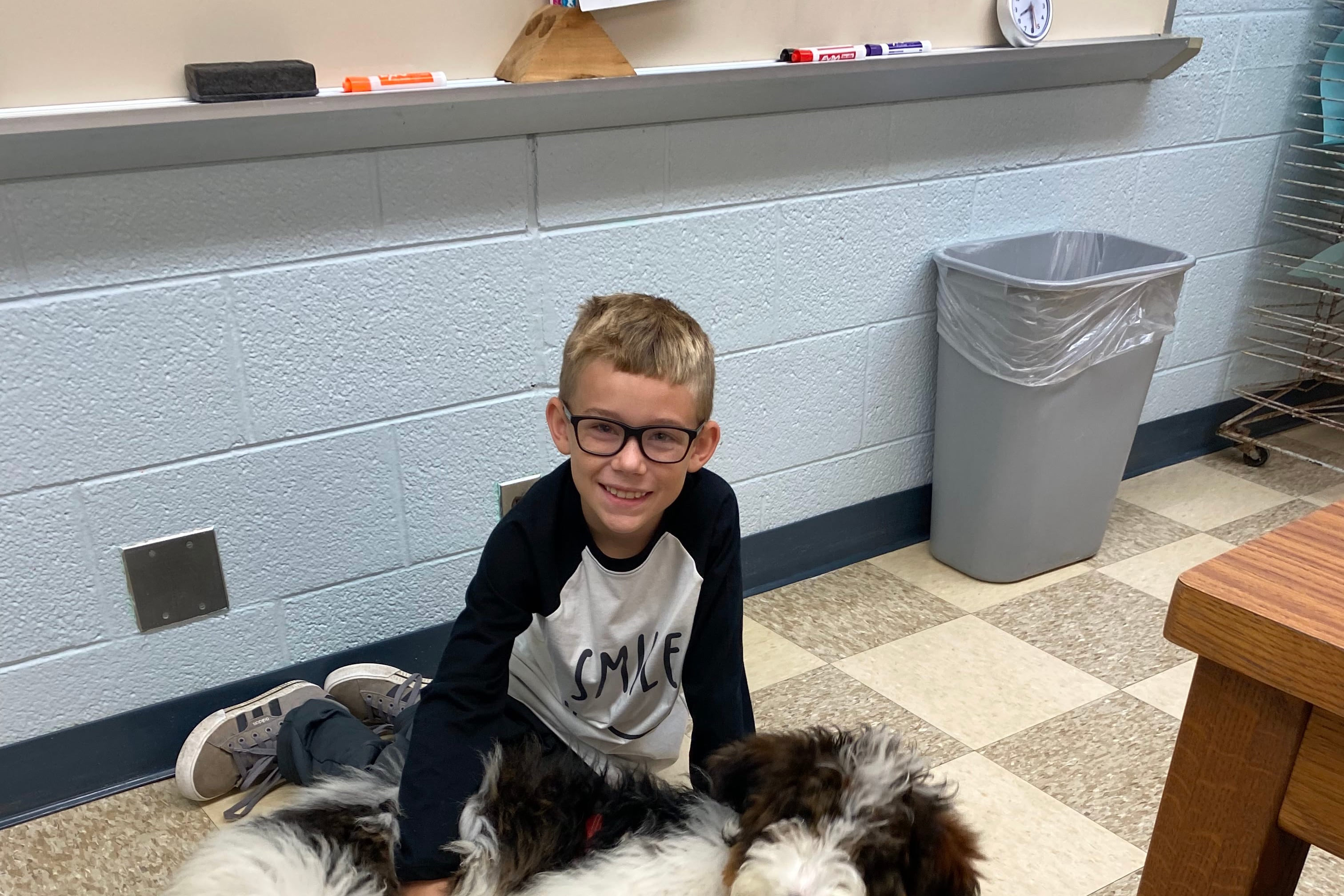 a student sits on the linoleum floor of a classroom with a hand on the dog sprawled in front of him