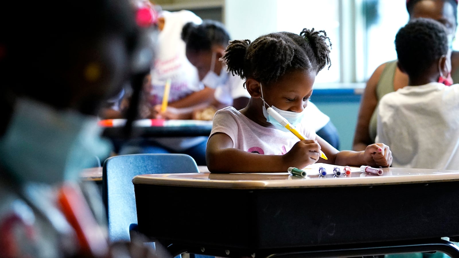A girl with pigtailed braids draws at her desk as other students work at theirs around her.
