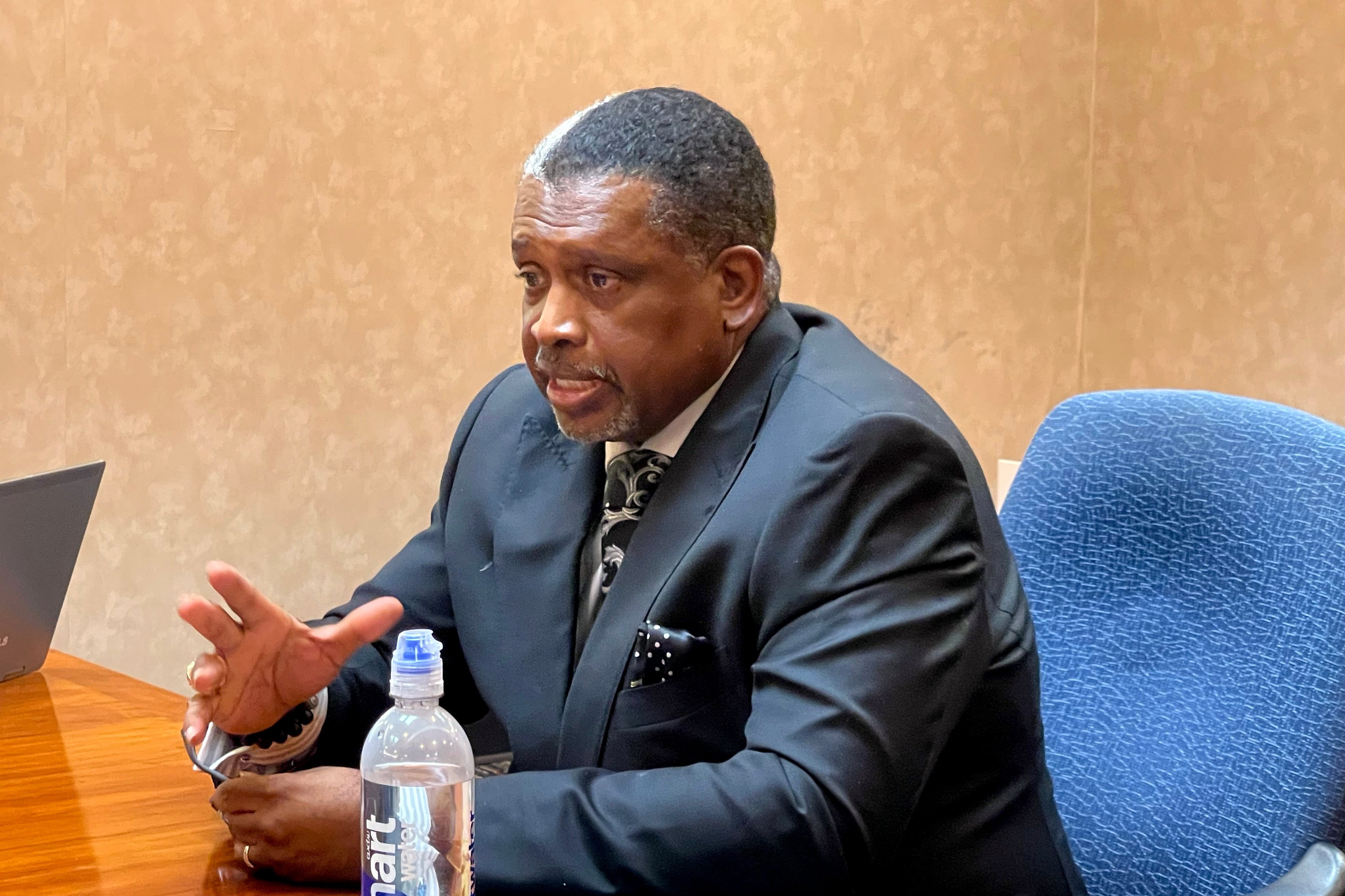 A photograph of a Black man in a suit sitting at a table with a water bottle in the foreground.