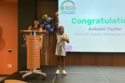 An adult stands behind a podium next to a young Black girl holding up a trophy and celebrating her award.