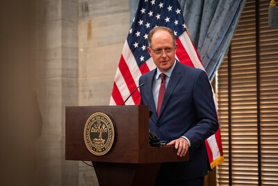 A man wearing a dark suit stands at a wooden podium with an American flag in the background.