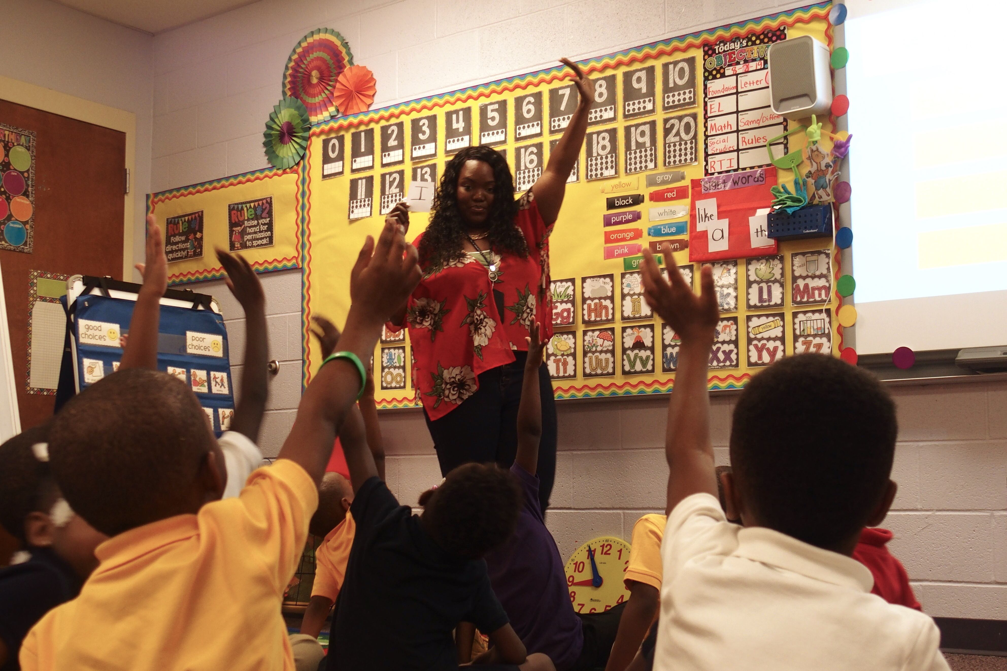 A Memphis teacher smiles and signals to her students sitting on the floor to raise their hands with her
