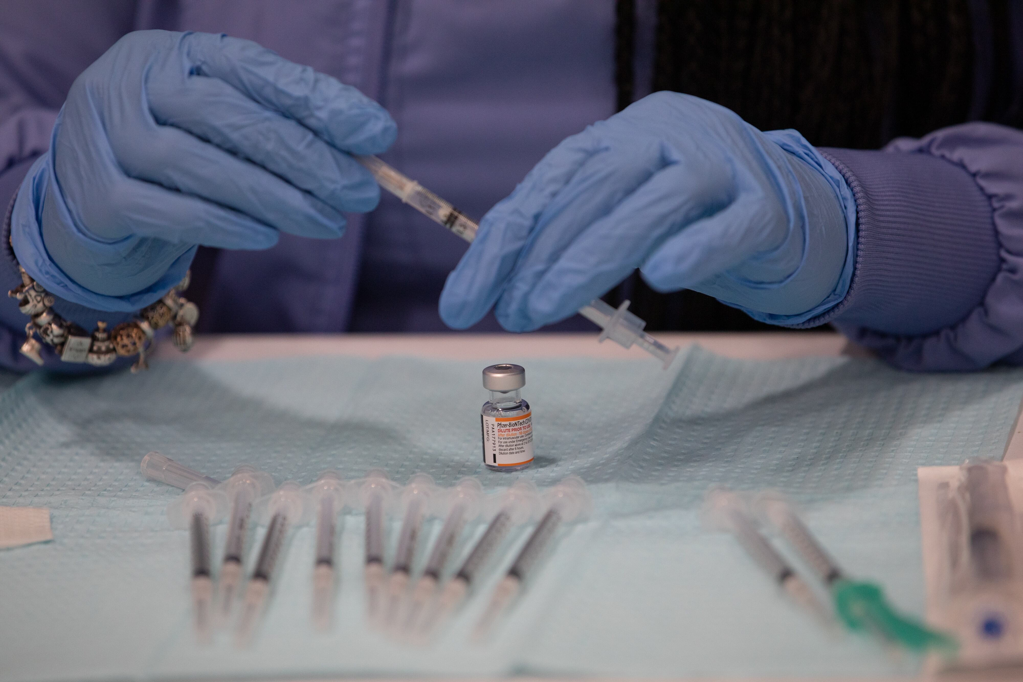 A close-up photo of a person’s hands wearing blue latex gloves, preparing a syringe for a COVID-19 vaccination.