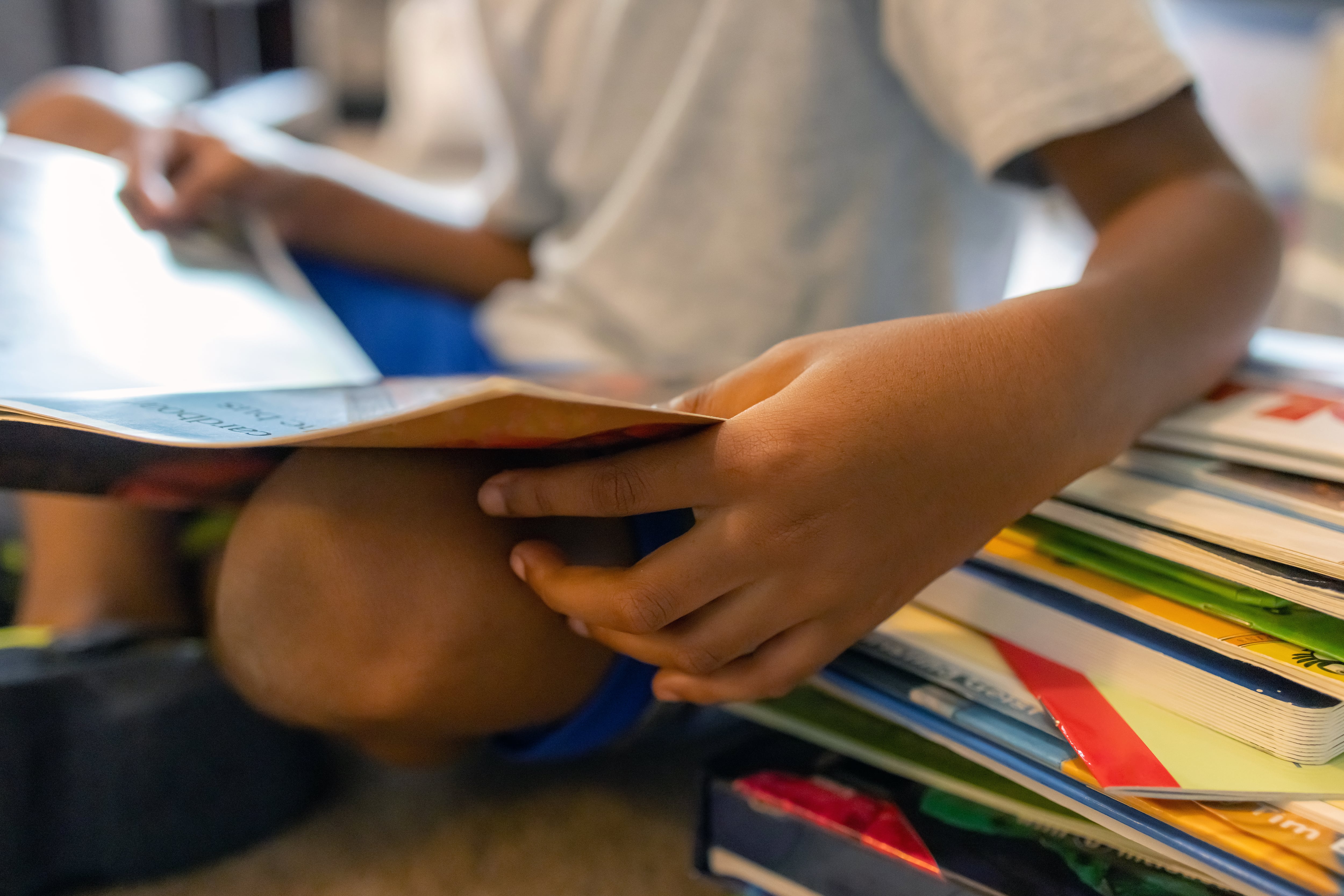 A non-identifiable young student sits on the floor reading a book next to a stack of books.