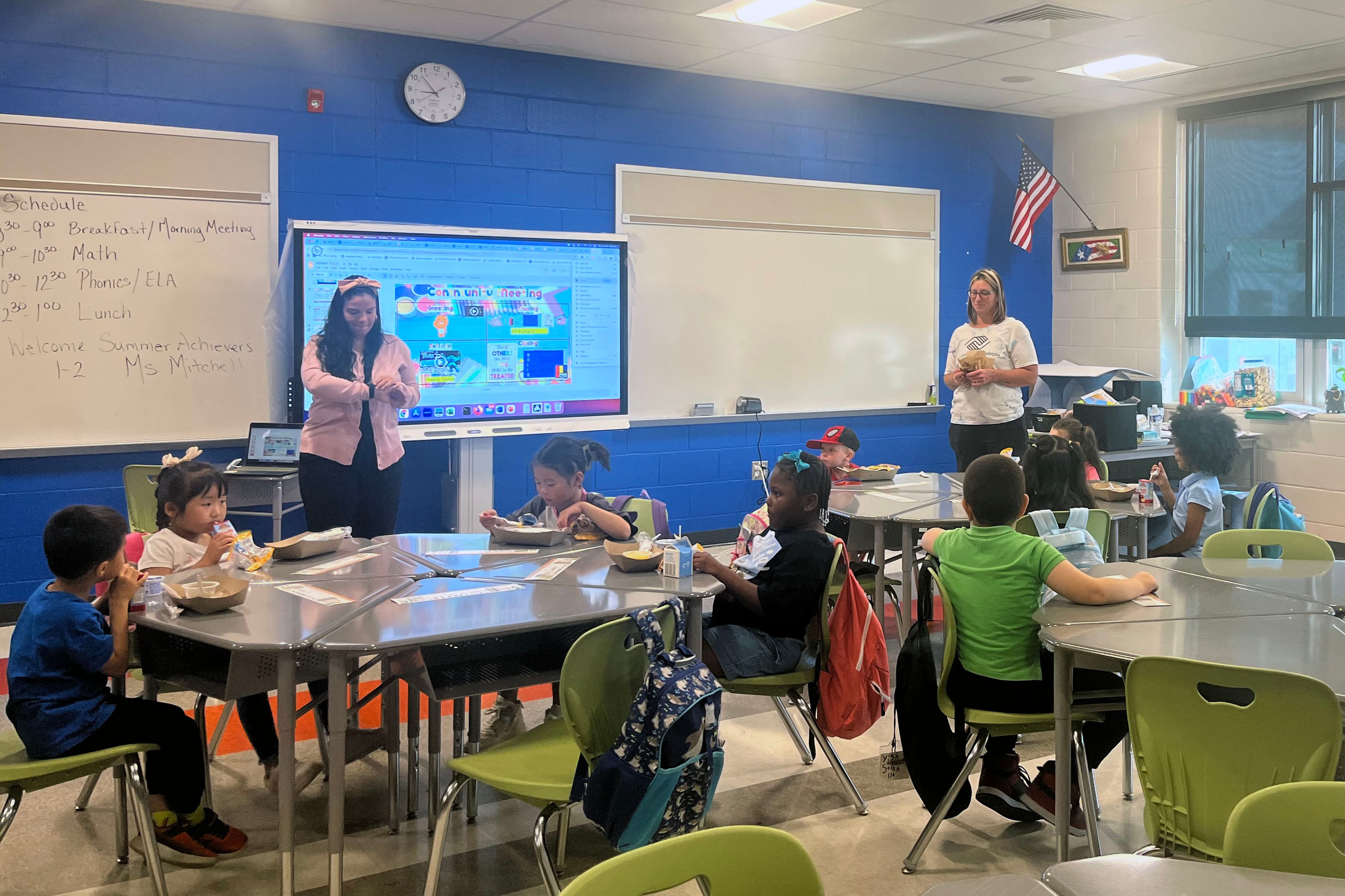 Two adults stand at the front of a classroom with a handful of young students sit at round tables.