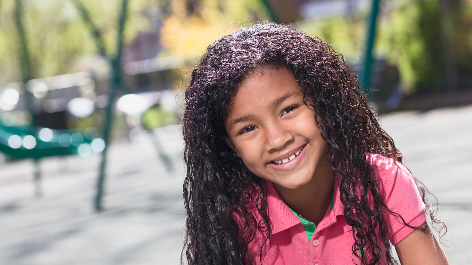 A little girl wearing a pink polo shirt smiles at a playground.