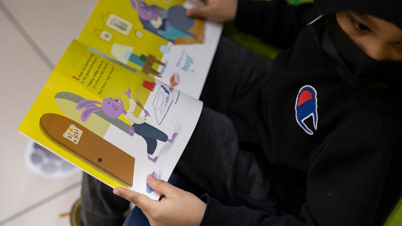A young boy in a black Champion hoodie and mask reads a children’s book.