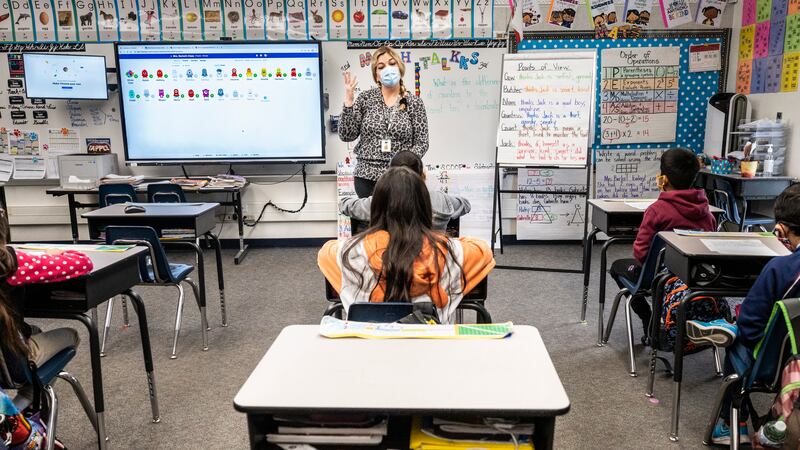 A teacher, wearing a blue face mask, stands in front of a whiteboard in a classroom. She is speaking to students who are also wearing masks and are seated at their desks.