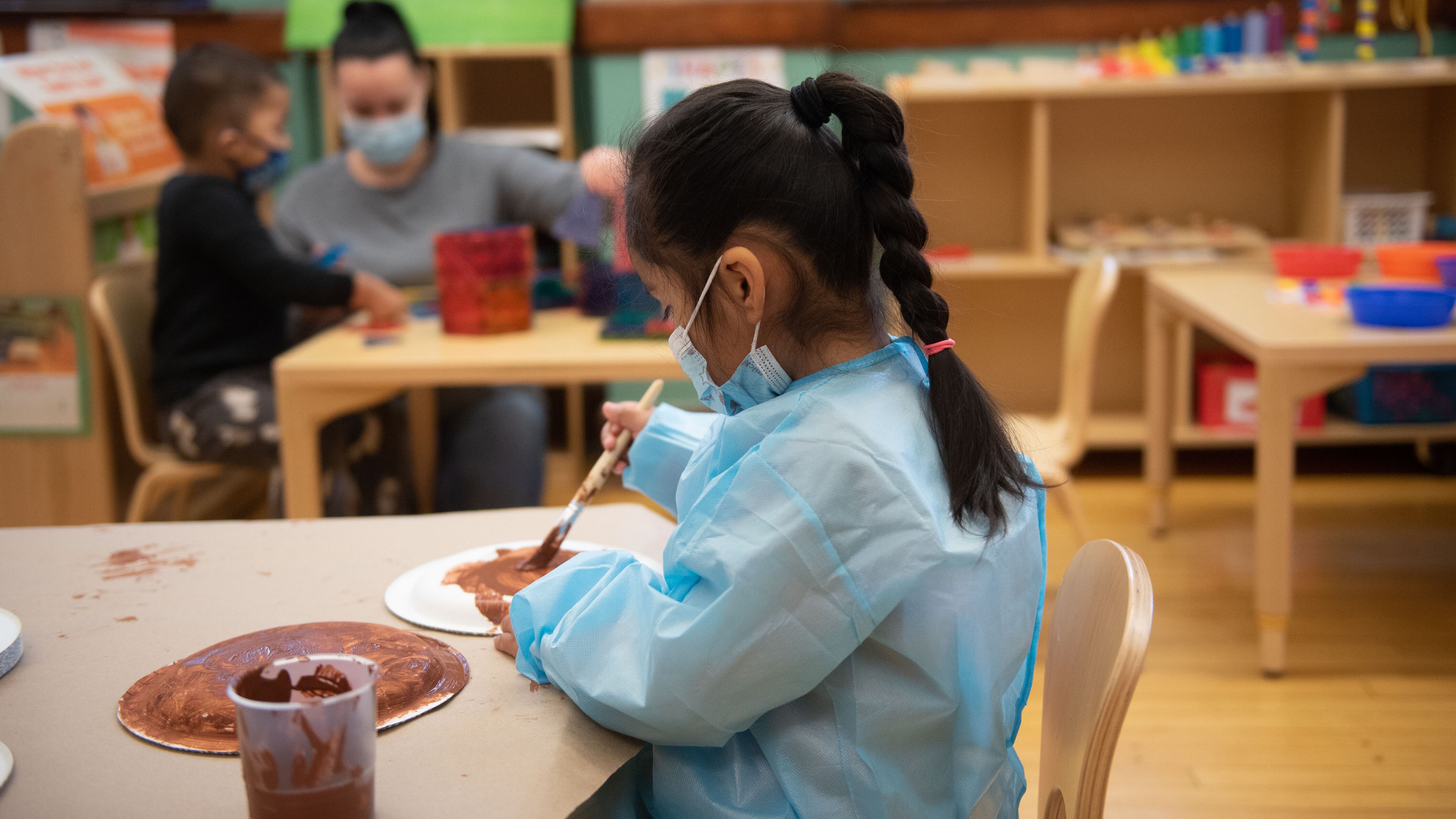 A student wearing a mask on her face and a blue smock to cover her clothes uses brown paint on paper plates.