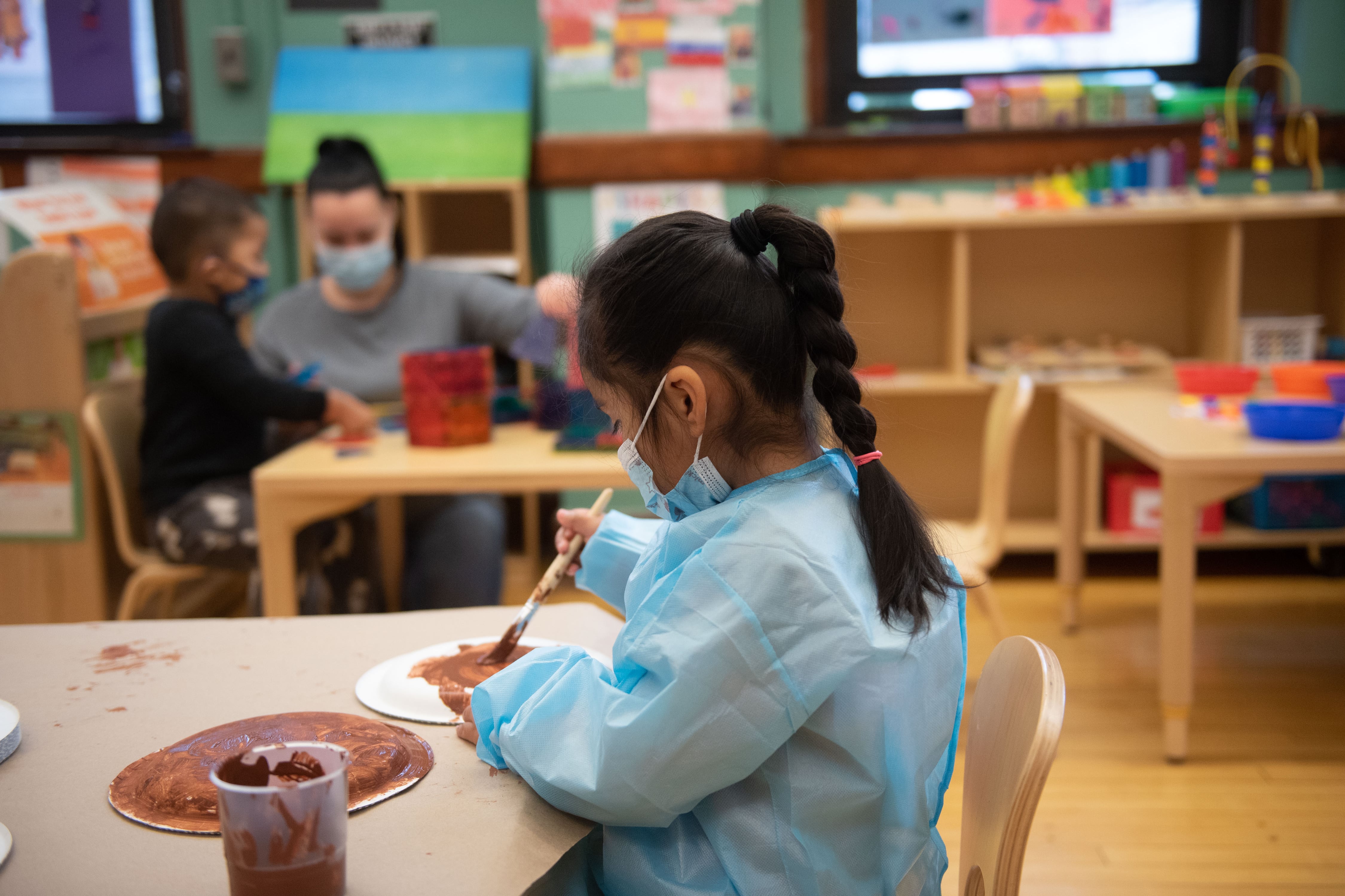 A student wearing a mask on her face and a blue smock to cover her clothes uses brown paint on paper plates.
