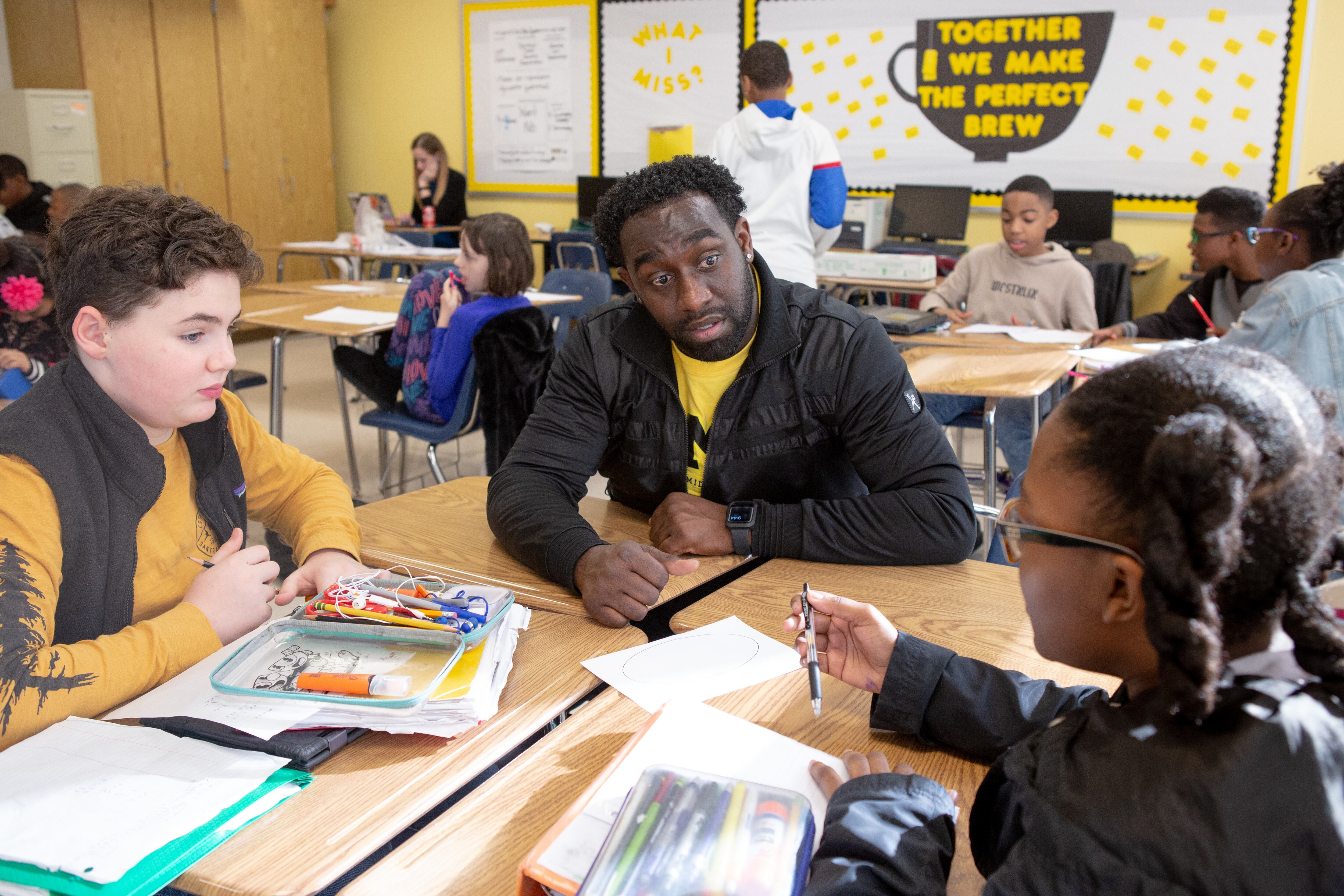 Two students, one a boy and a girl, are sitting with a teacher in a black jacket and yellow shirt. In the background, students are sitting at desk appearing to do a classroom assignment.