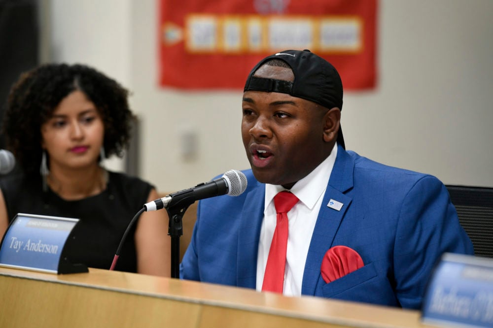 Tay Anderson, wearing a blue suit, red tie and handkerchief, and black baseball hat, speaks into a microphone from the dais of the Denver school board.