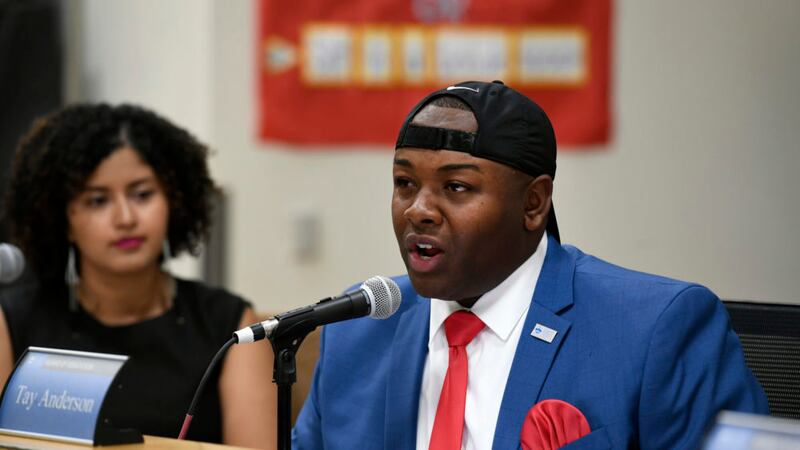 Tay Anderson, wearing a blue suit, red tie and handkerchief, and black baseball hat, speaks into a microphone from the dais of the Denver school board.