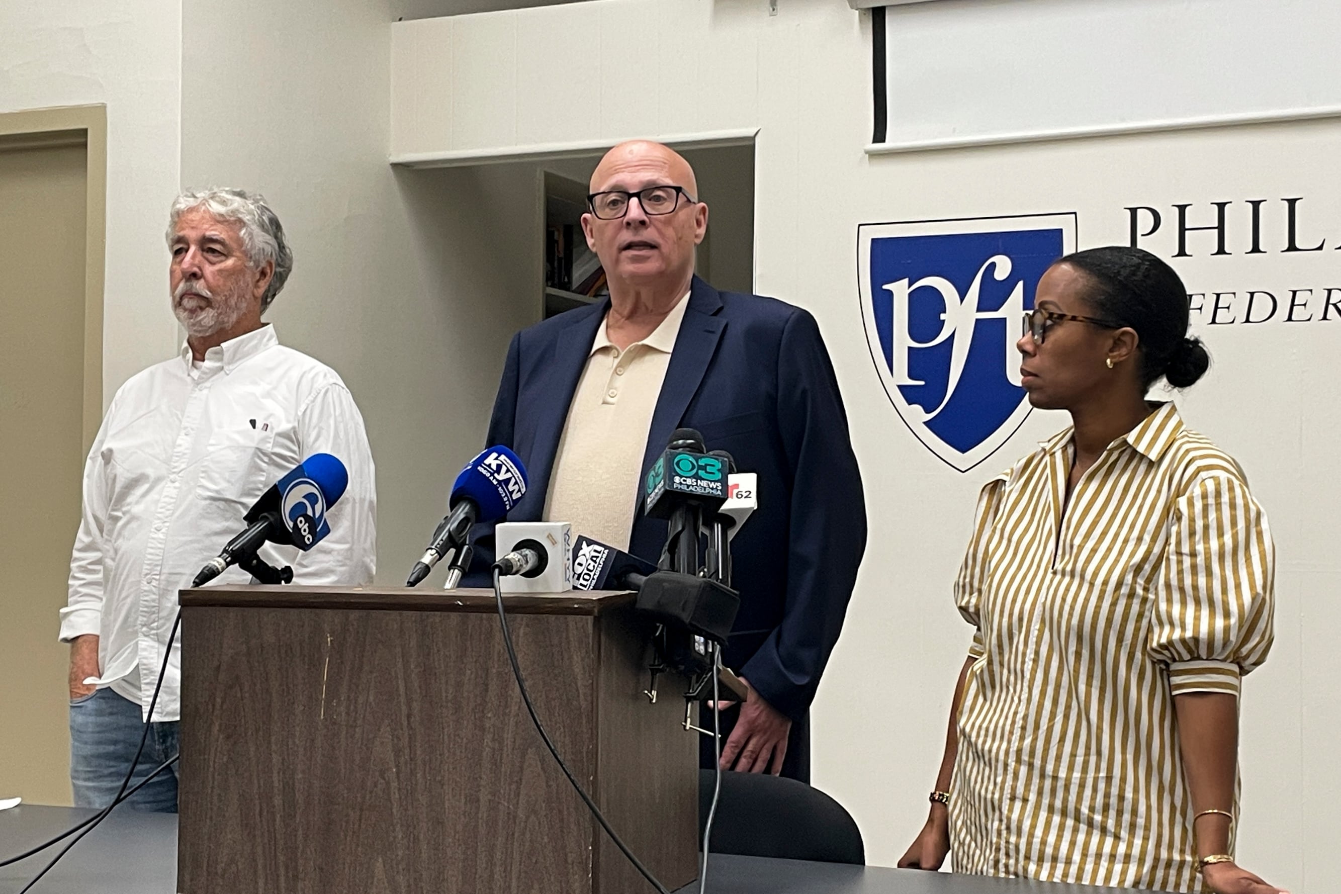 A man wearing glasses is speaking at a podium, with two other people standing beside him on each side.
