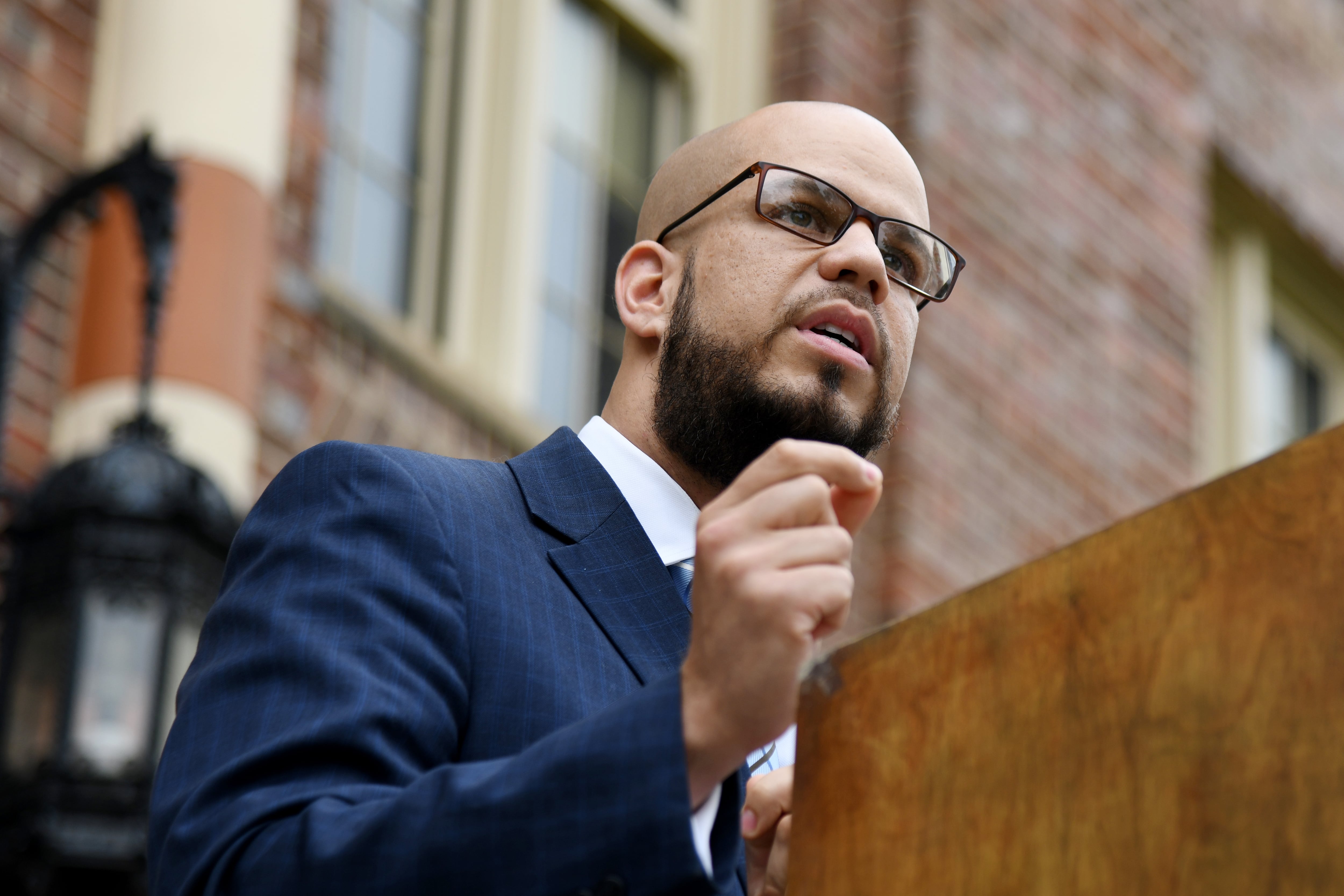 Denver Public Schools Superintendent Alex Marrero, wearing glasses and a blue checkered suit jacket, makes a speech outdoors at a wooden podium.