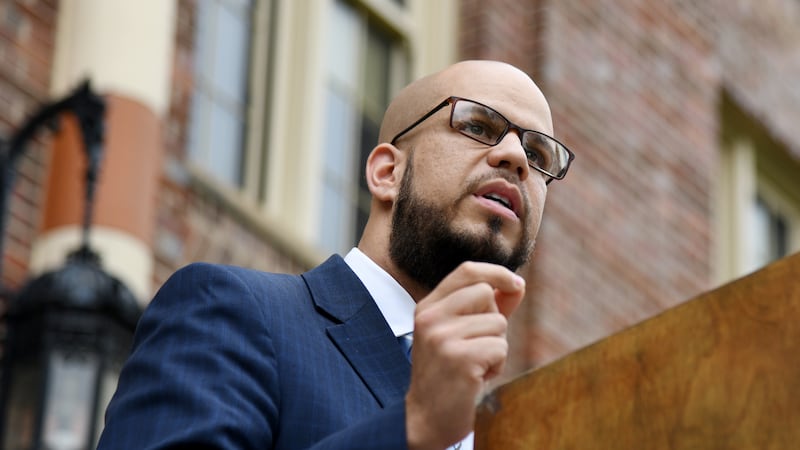 Denver Public Schools Superintendent Alex Marrero, wearing glasses and a blue checkered suit jacket, makes a speech outdoors at a wooden podium.