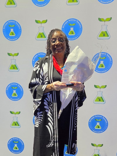 A photograph of a Black woman holding a bouquet of flowers and smiling for a portrait with a banner in the background.