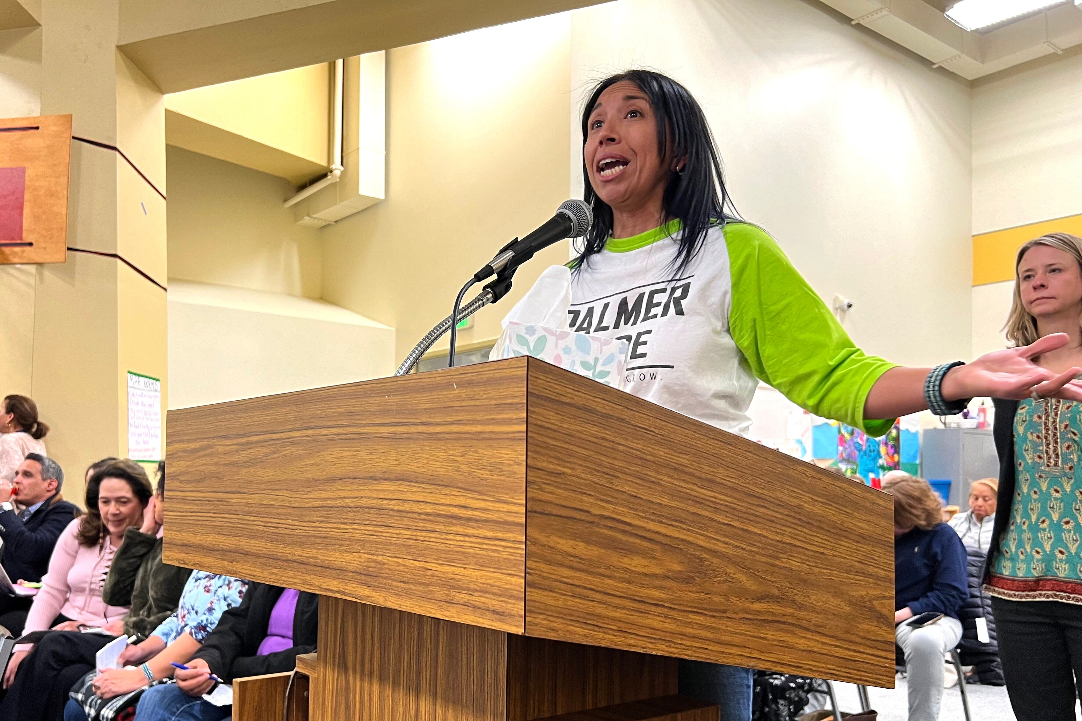 A woman in a green-and-white top stands at a podium and gestures while speaking.