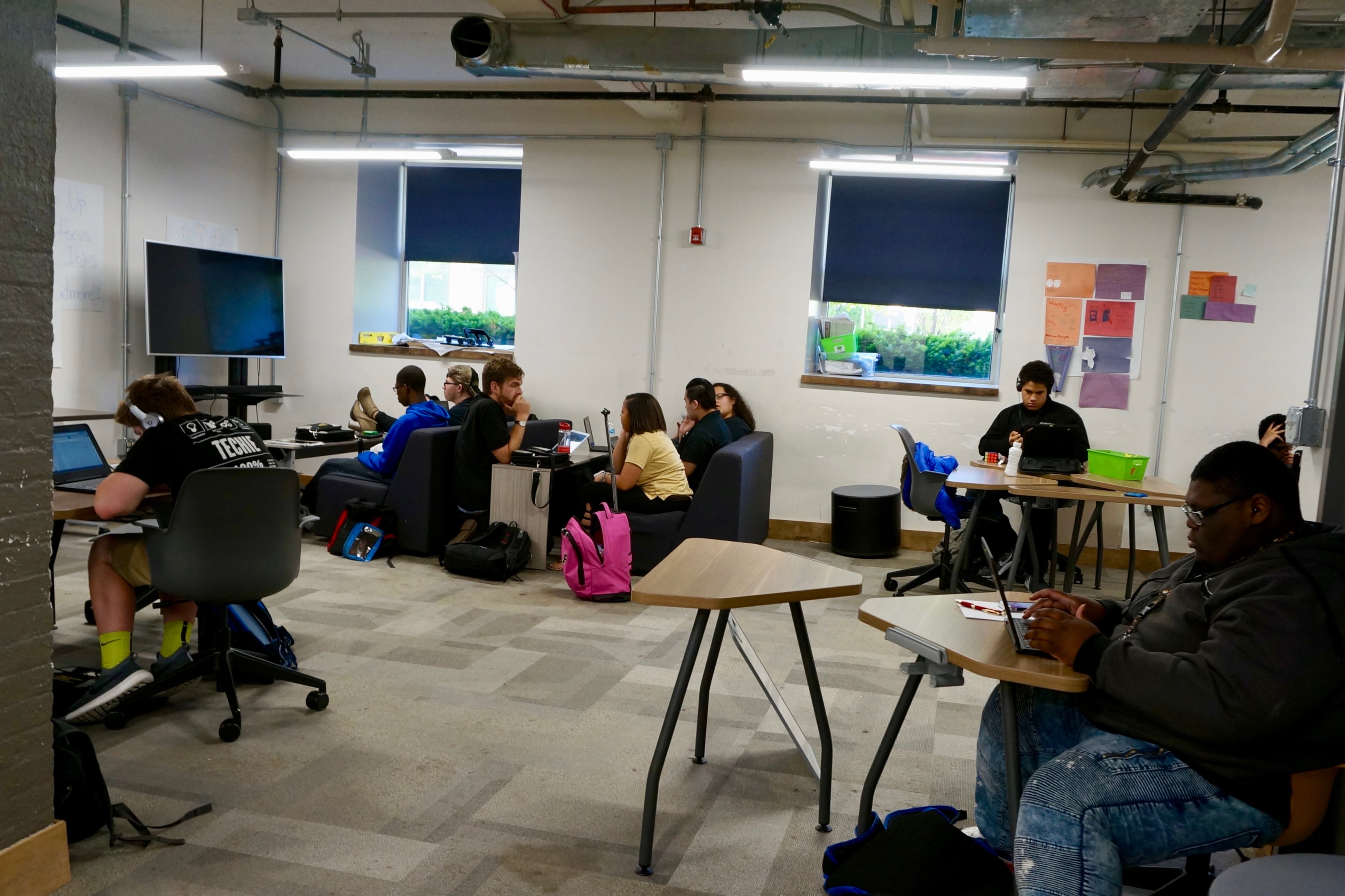 Students sit in a classroom, each one working at a desk. Some have headphones on. Others are on laptops.