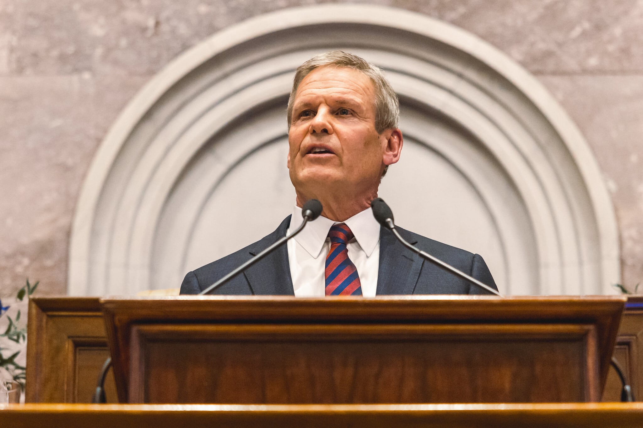 A man in a suit speaks from behind a wooden podium with two microphones and in front of a light background.