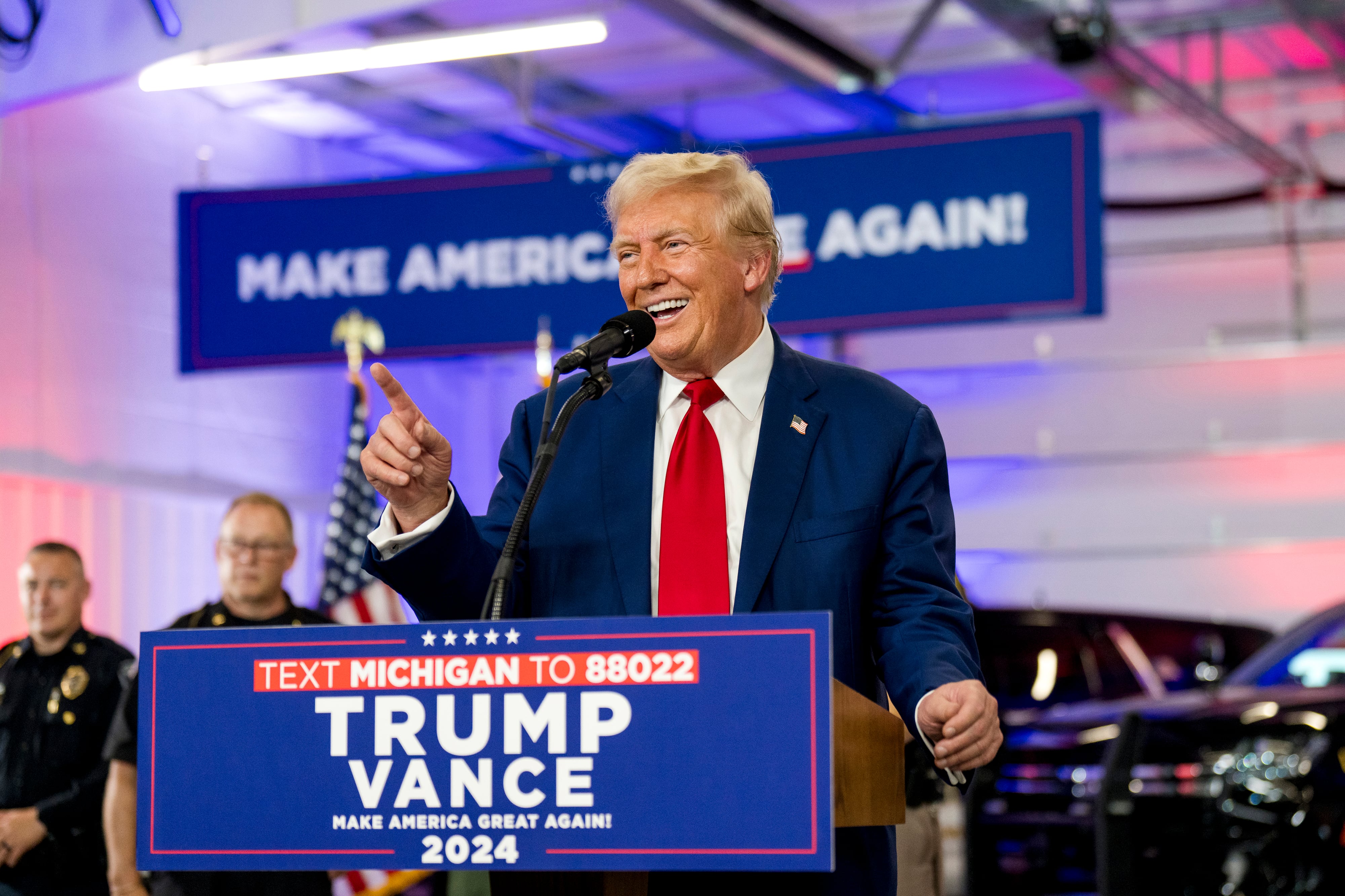 A man wearing a dark suit and a red tie stands behind a podium and in front of a large projected sign that reads "Make America Great Again!"