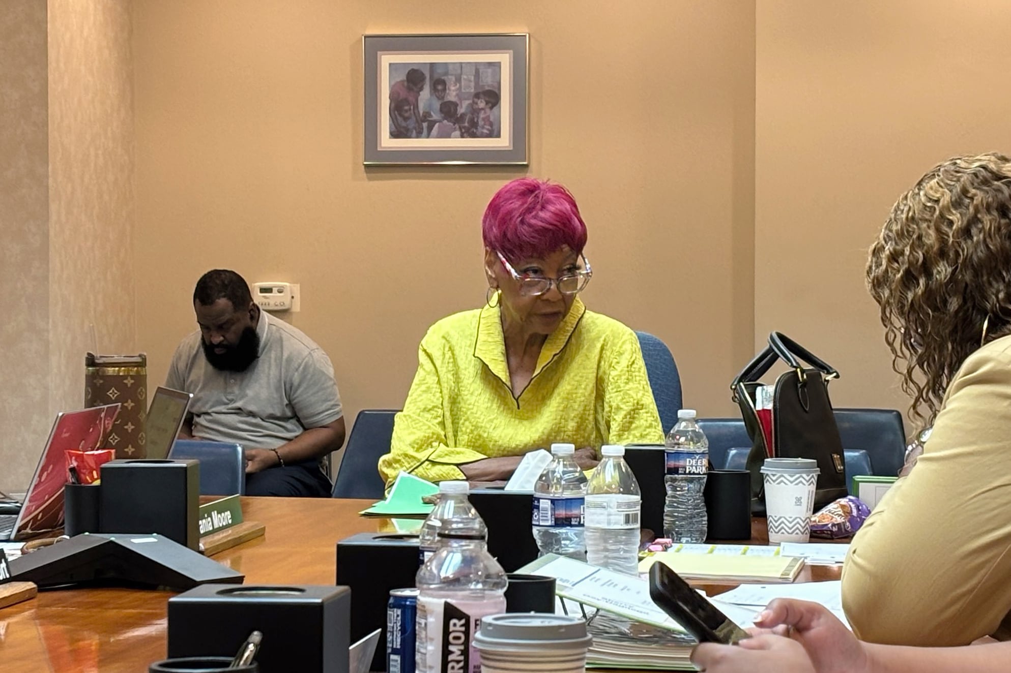 A photograph of a woman with short red hair and wearing a yellow top sits at a wooden table with one person on each side.