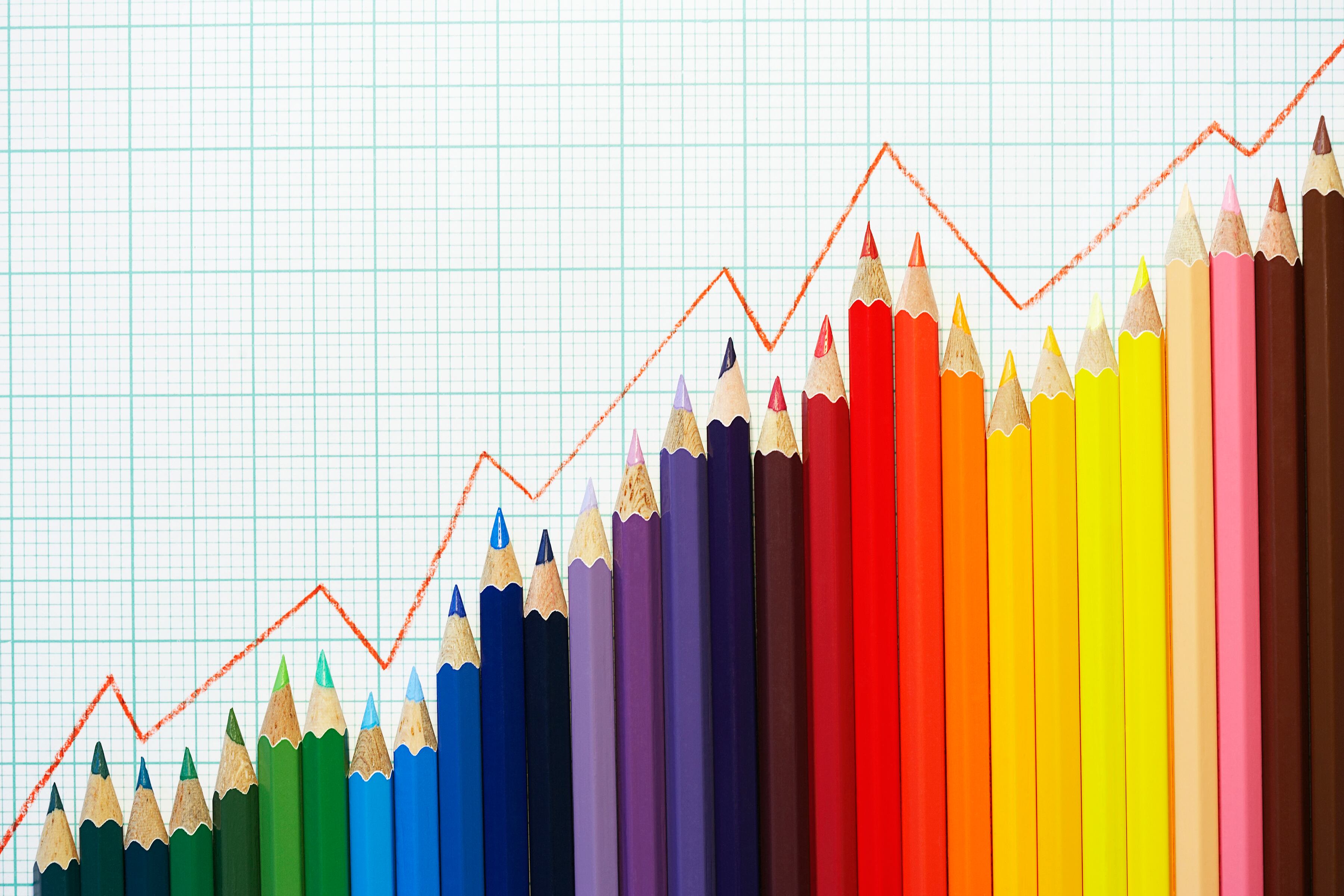 Colored pencils are lined up in the shape of a graph with a read line following the top of the pencil tips with white and blue graph paper as the background.