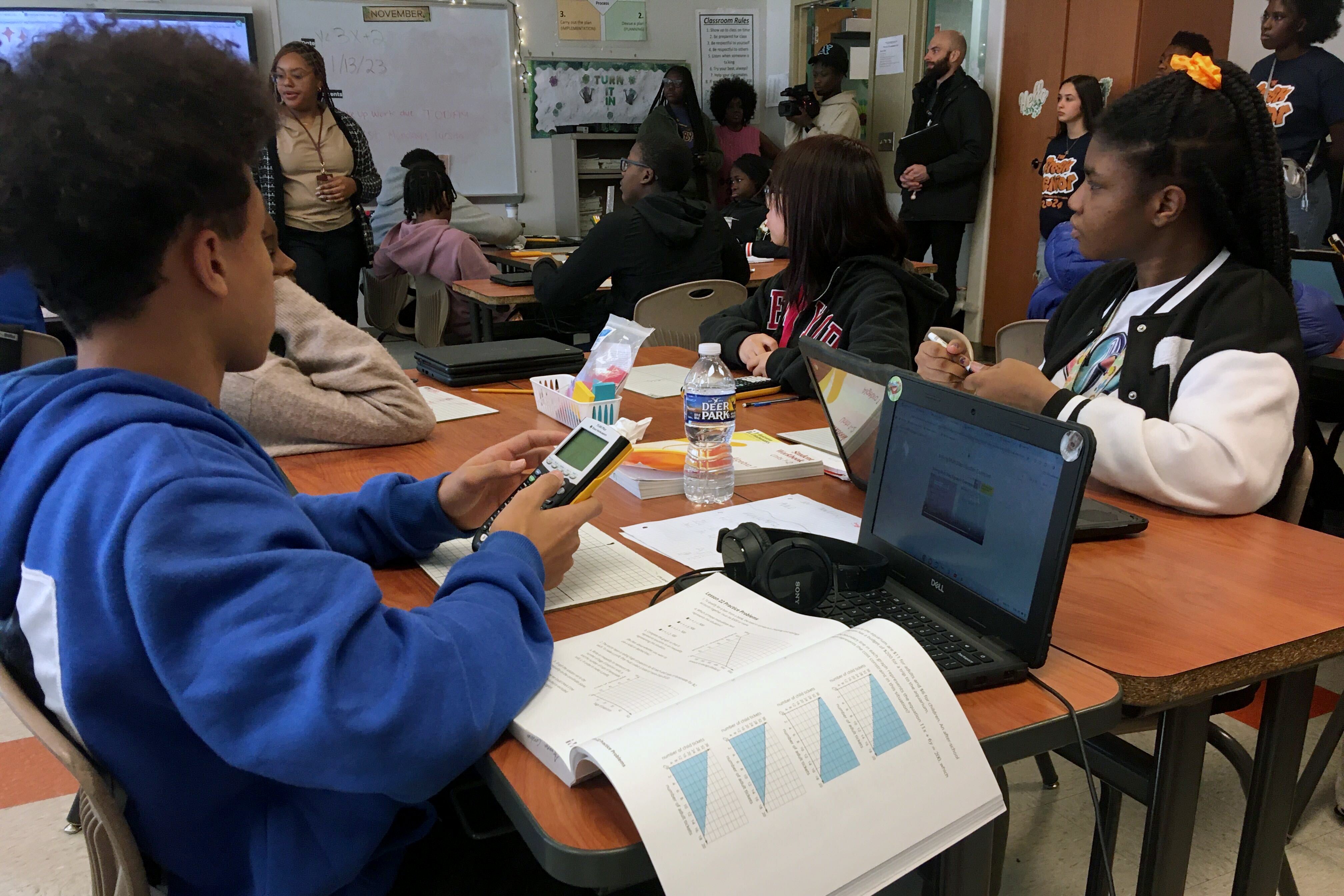 Multiple students work at desks in a classroom with adults and a teacher standing along the wall in the background.