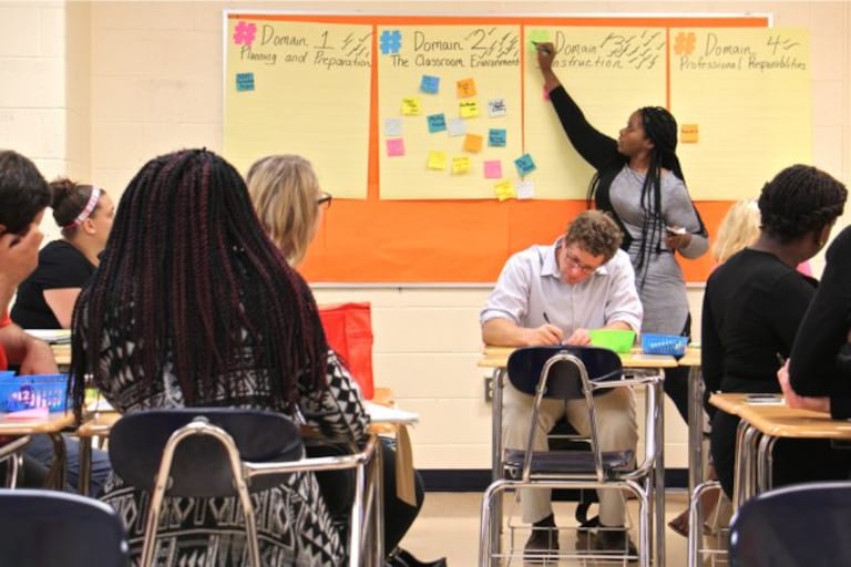 A woman points to yellow posters with Post-It notes on the wall as adults sitting at desks look on.