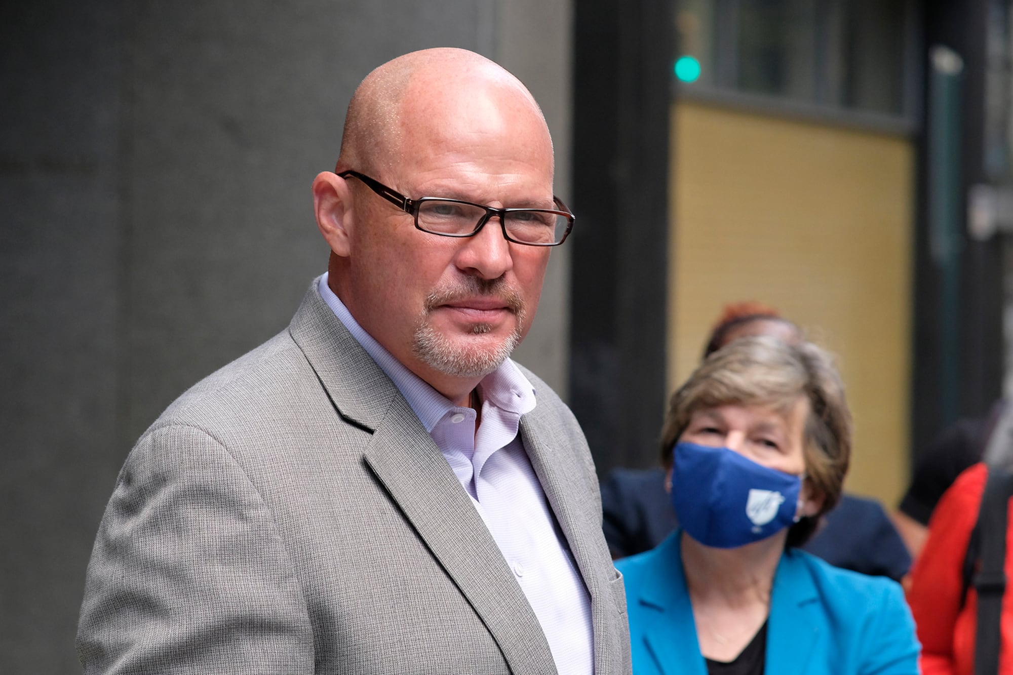 A photograph of a white man wearing glasses and a grey suit outside with a person wearing a blue face mask in the background.
