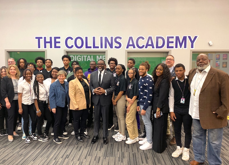 A group of students, staff and the Chicago Mayor Johnson, line up and pose for a portrait in front of a white wall with windows and a sign that reads "The Collins Academy" on the top.