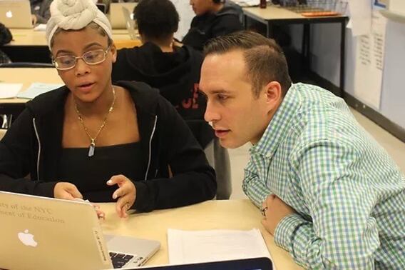 A teacher in a checkered shirt looks at a student’s work while speaking to her at a desk.