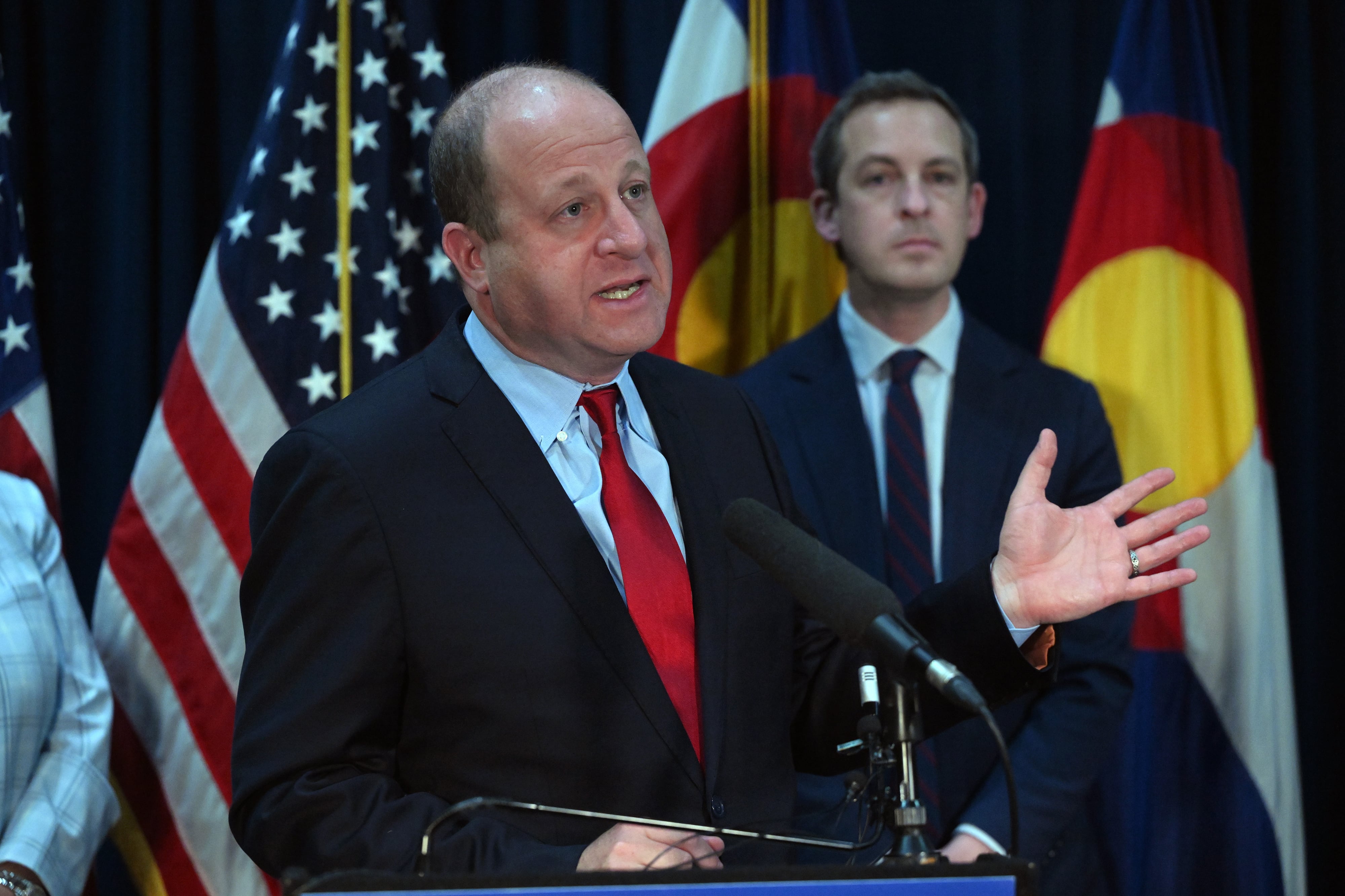A man speaks from a podium while another man stands in the background by an American and Colorado state flags.
