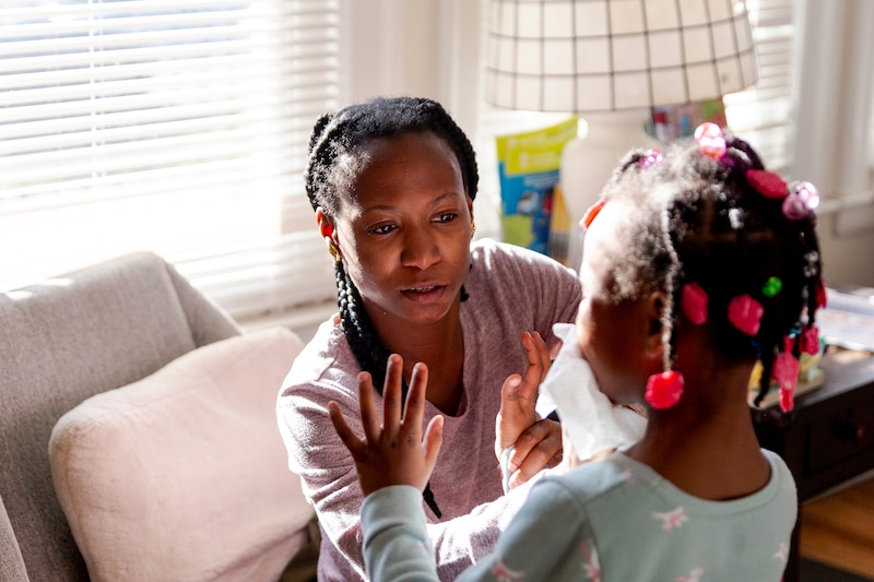 A woman sitting in a chair in a living room next to a large window is helping take care of a young child with colorful beads in her hair.