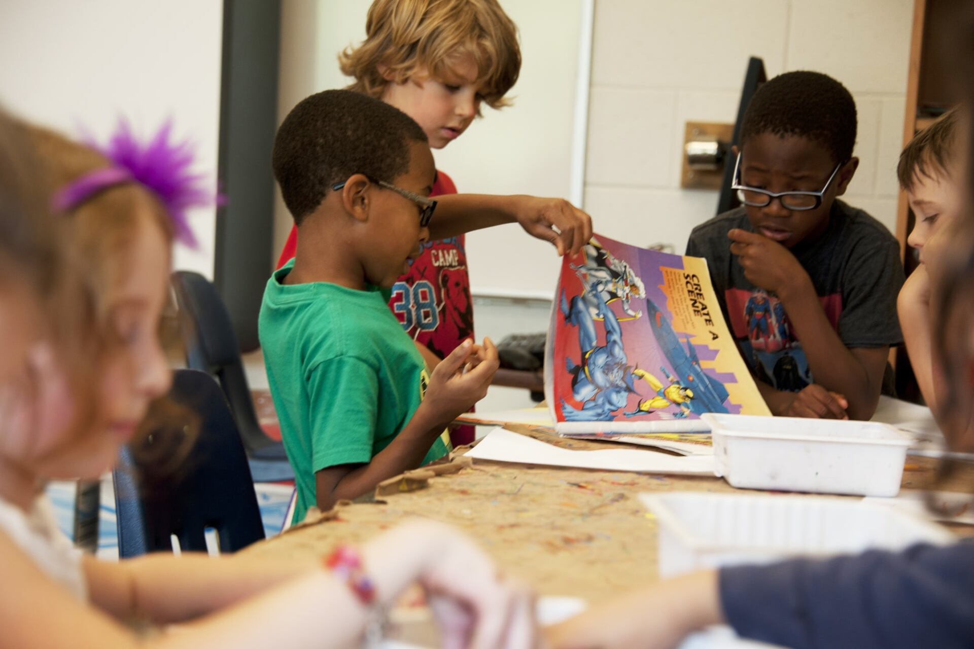 A group of four schoolboys gathered at a brown paper-covered table, studying a stylized cartoon book to be used as reference material, for a drawing they were asked to create in the classroom.