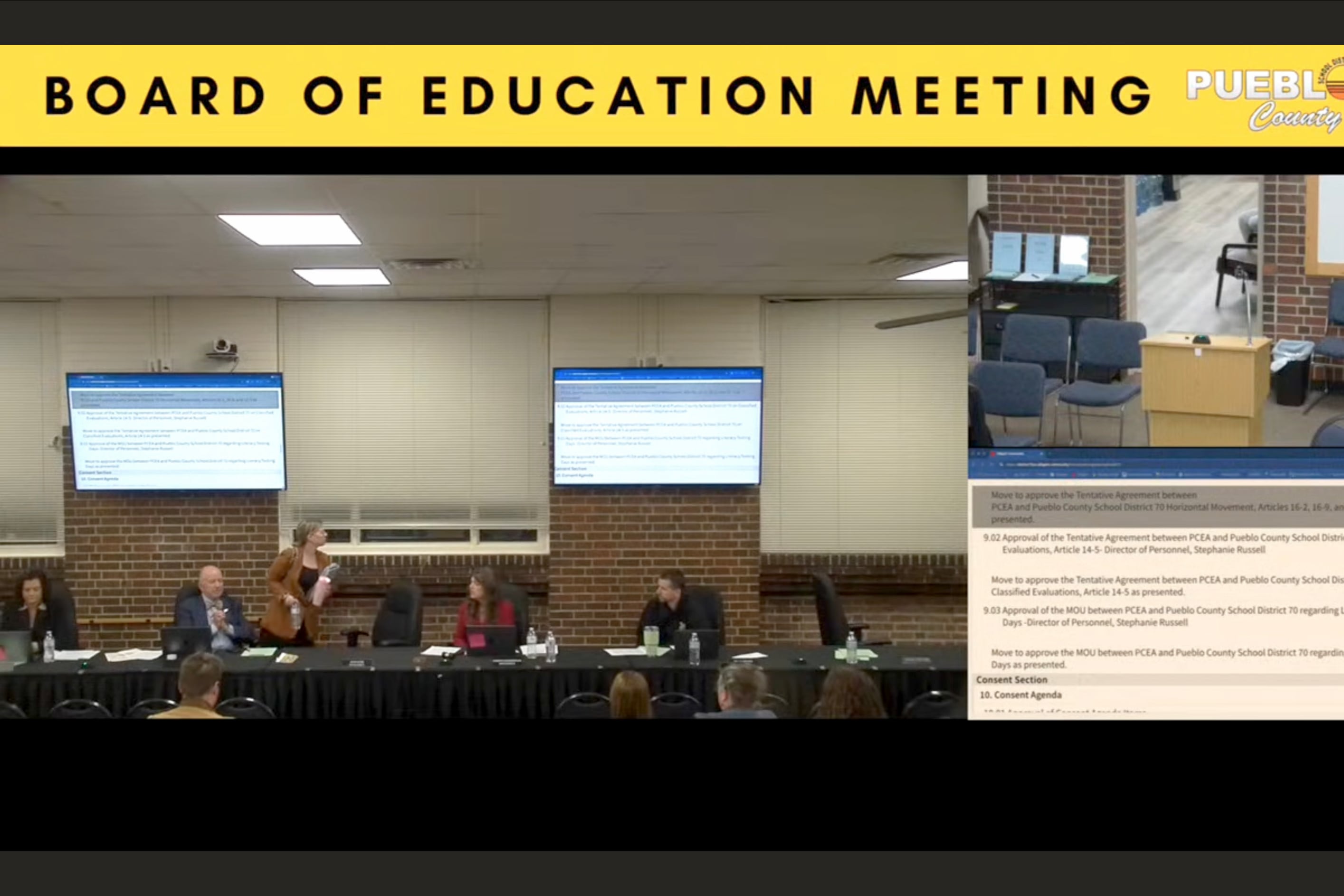 A screengrab of a group of people sitting at a long table in a large meeting room.