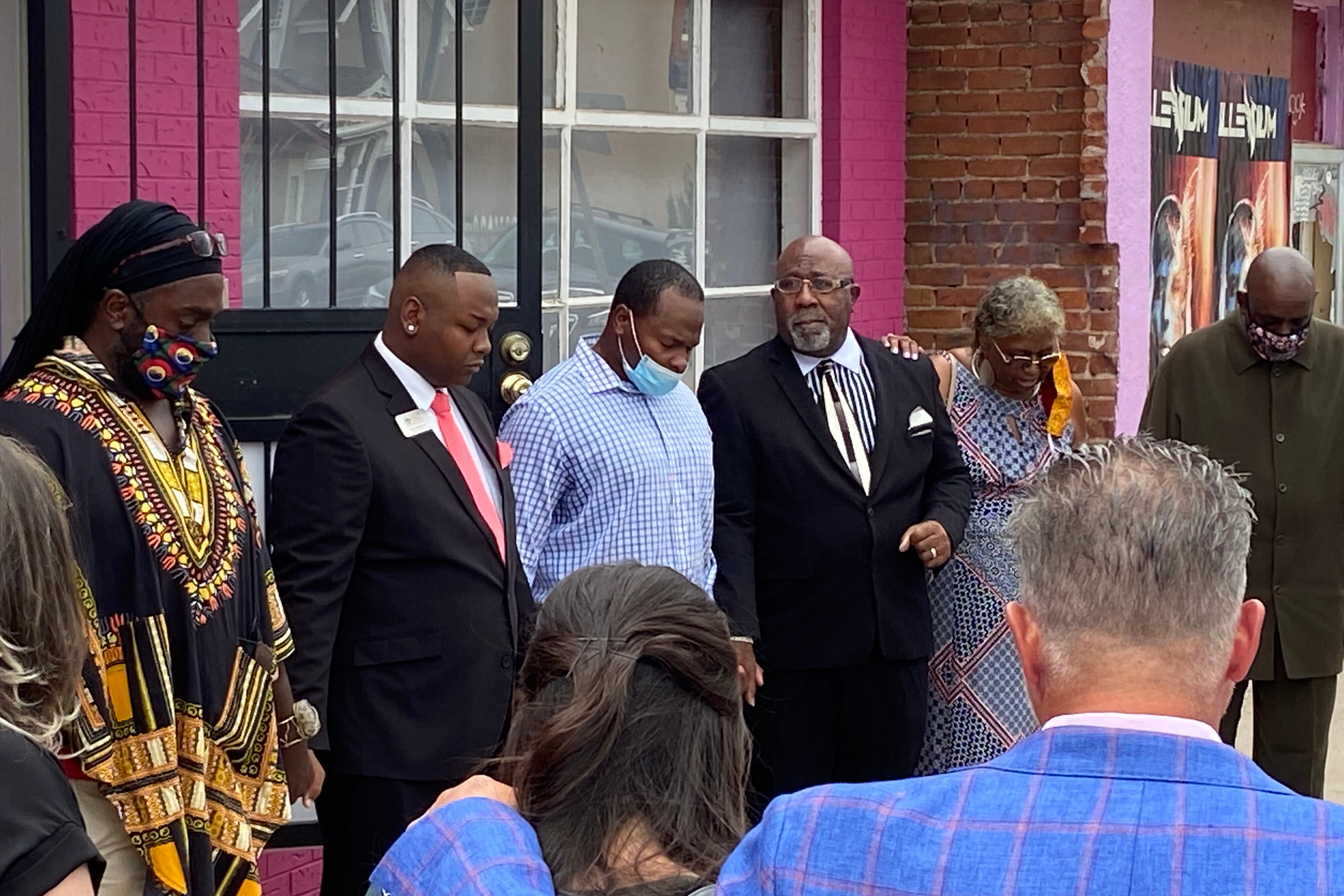 Denver school board member Tay Anderson, second from left, dressed in a suit and red tie, stands with his head bowed in prayer. He is standing in a circle of supporters holding hands.