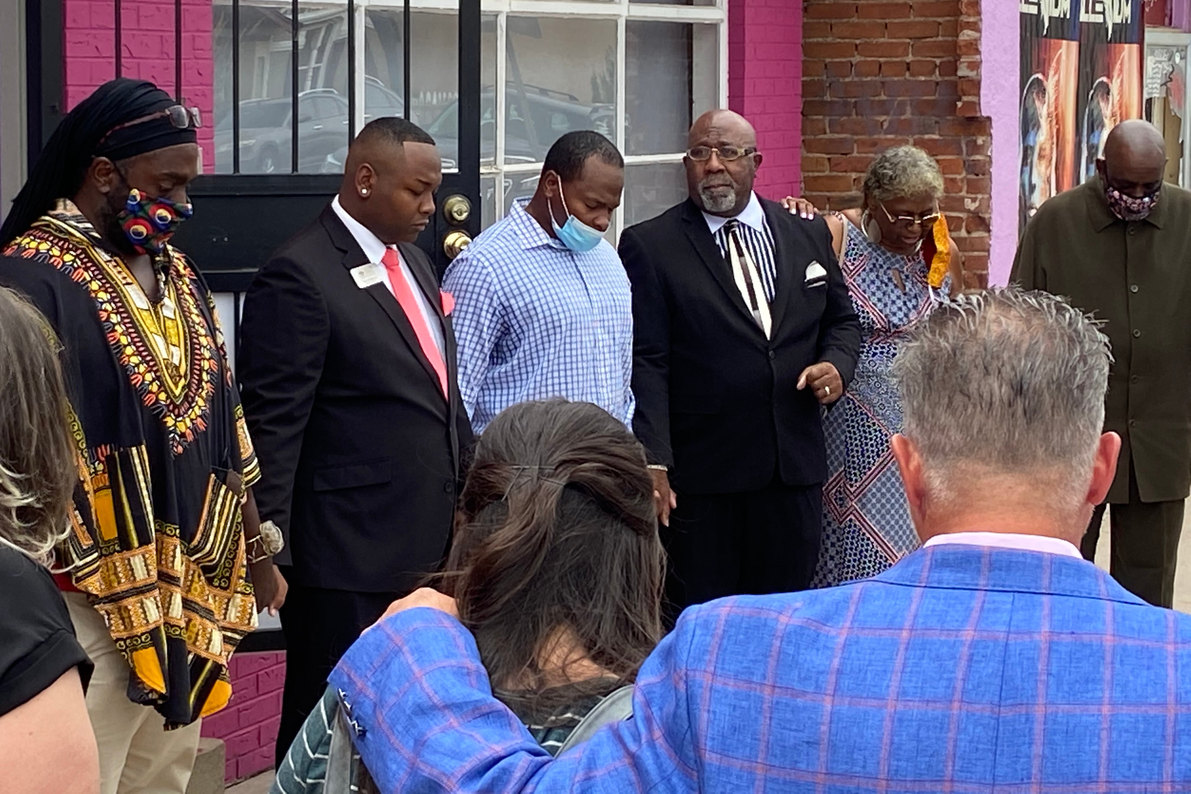 Denver school board member Tay Anderson, second from left, dressed in a suit and red tie, stands with his head bowed in prayer. He is standing in a circle of supporters holding hands.