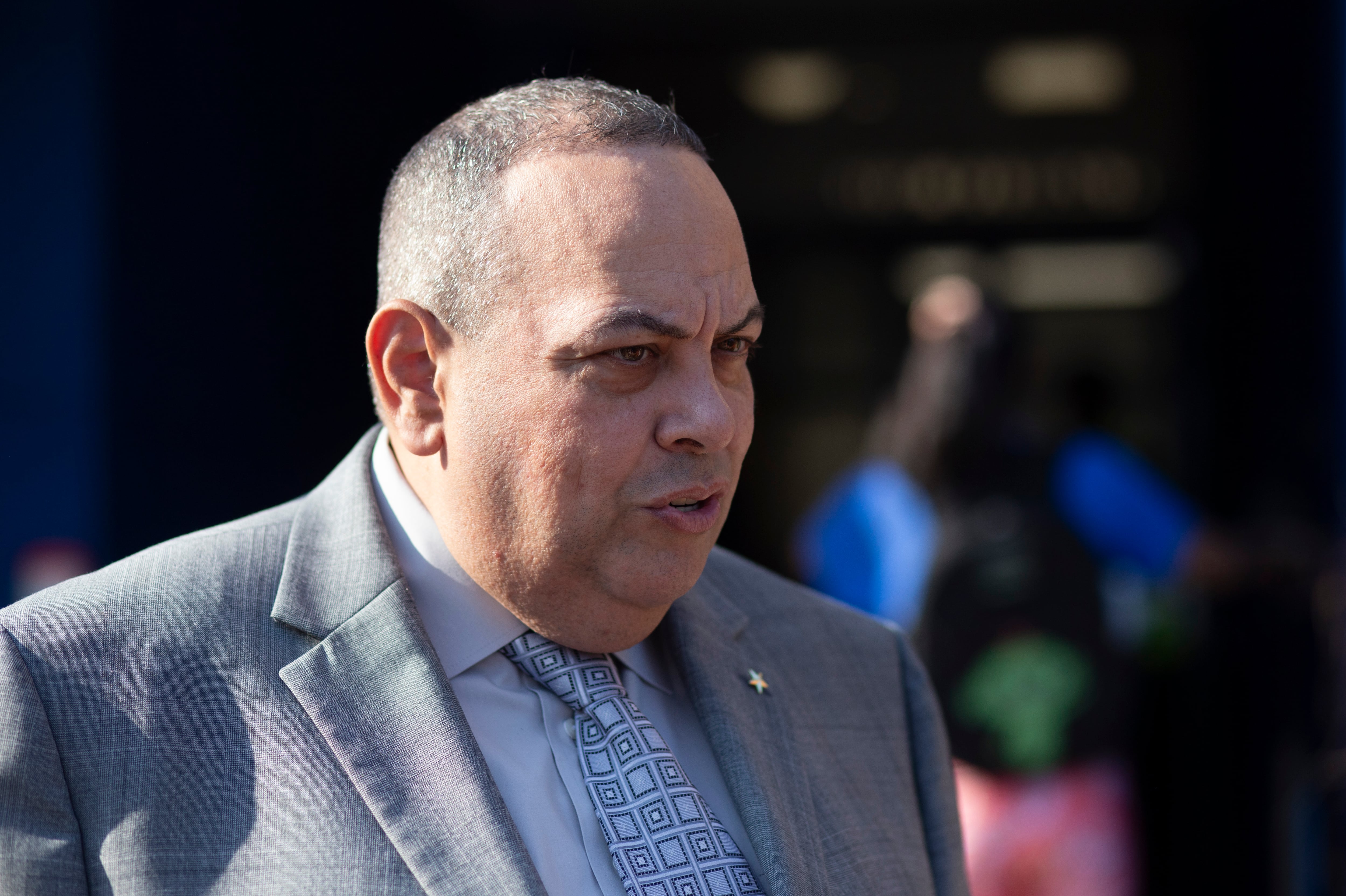 A man with short hair and wearing a light suit stands outside.