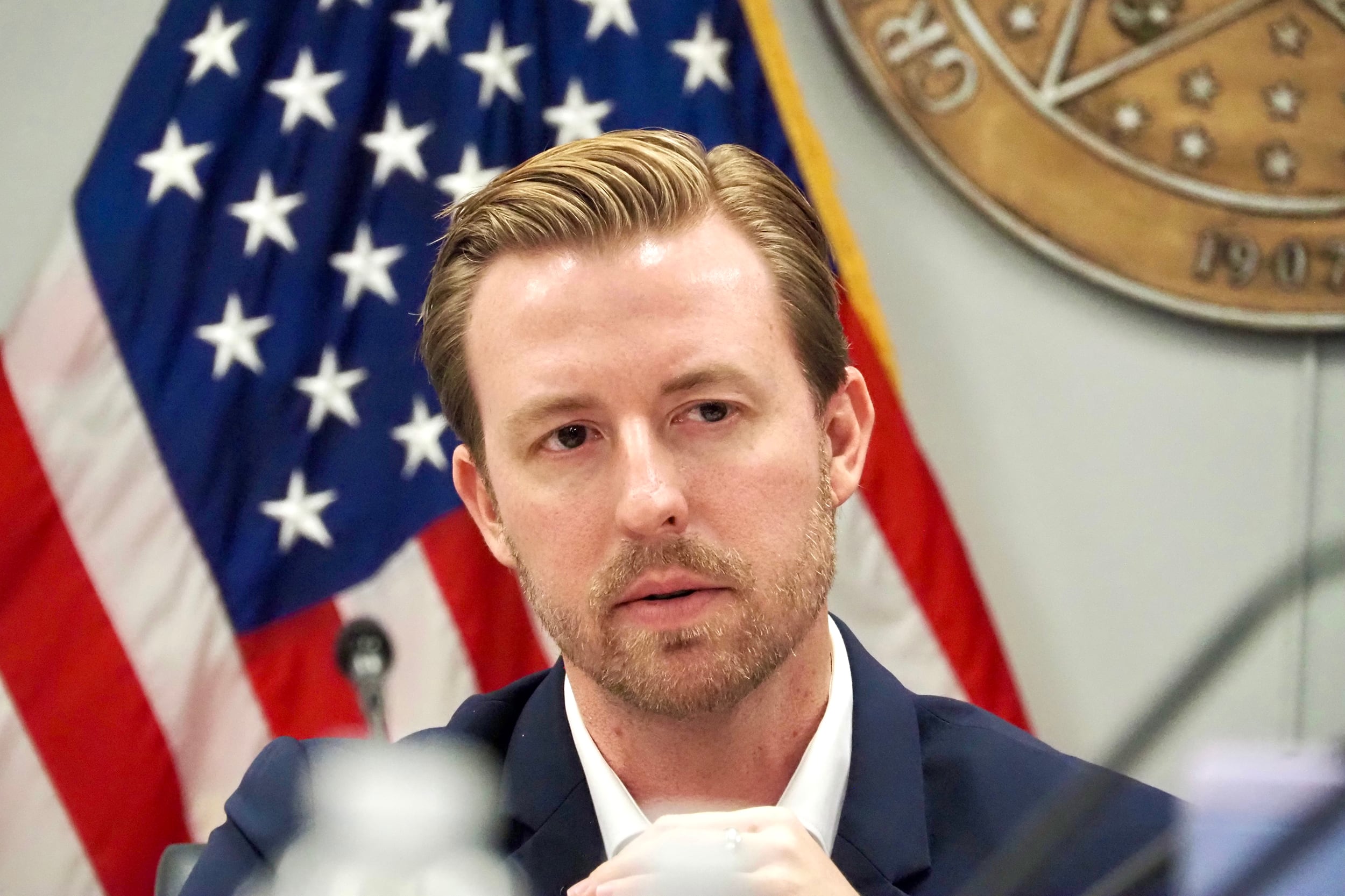 A photograph of a white man sitting in front of an American flag.