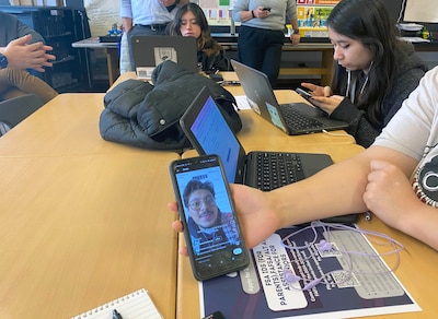 A hand holds out a phone with three students in the background all sitting at a large wooden table.