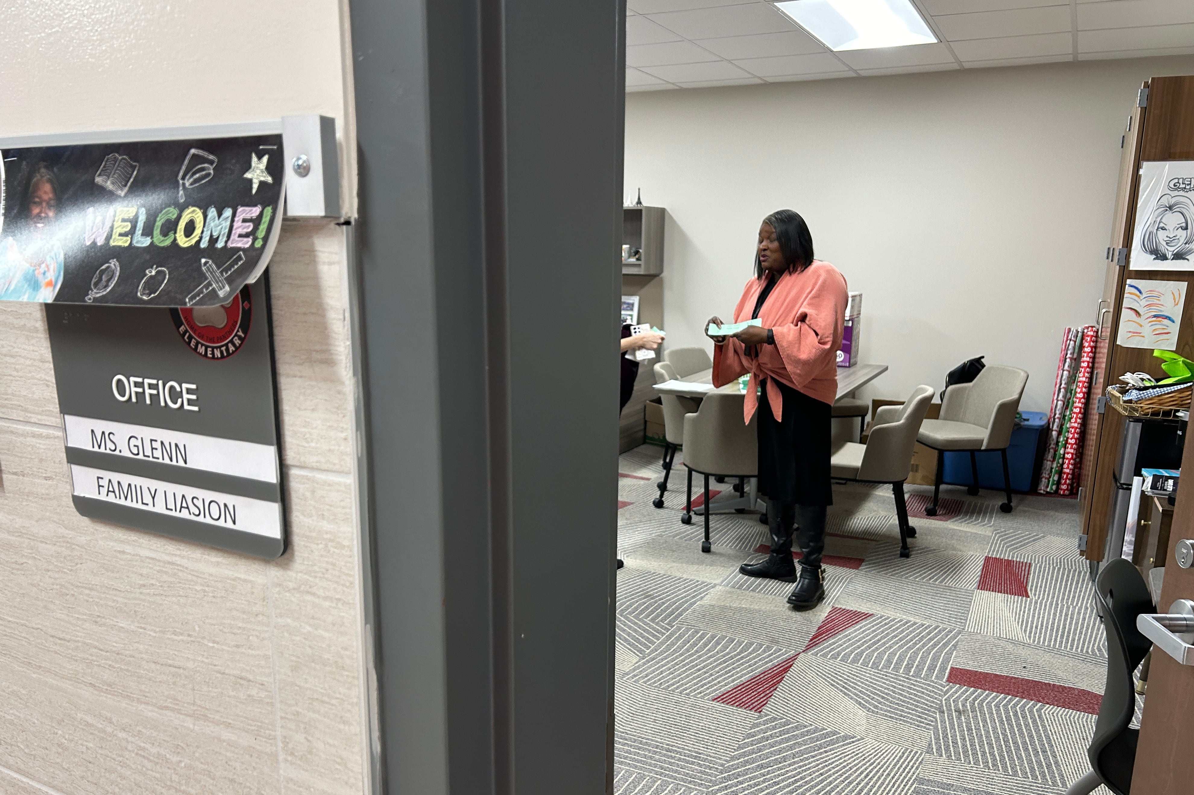 A woman with medium length dark hair and wearing a light pink sweater stands in her office with chairs in the background. On the left, there is a welcome sign and directory plate on the wall.