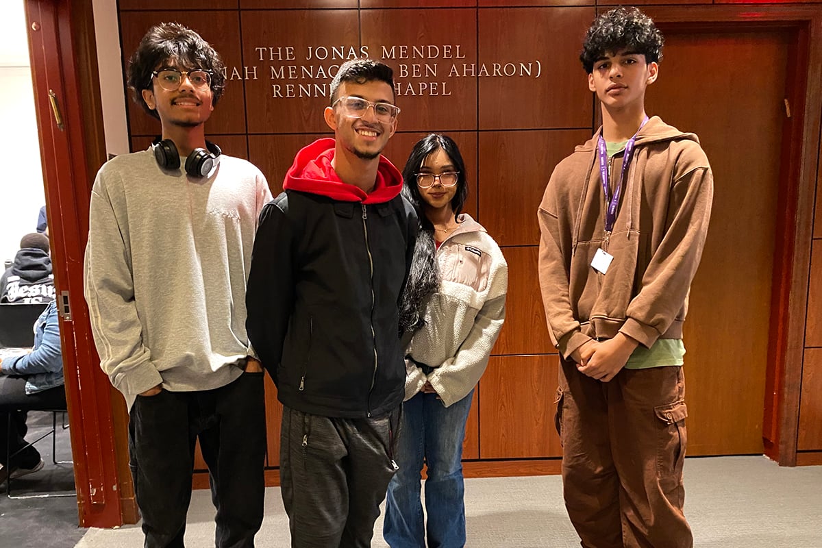 Four teenagers in casual clothes stand together in front of a dark wall.